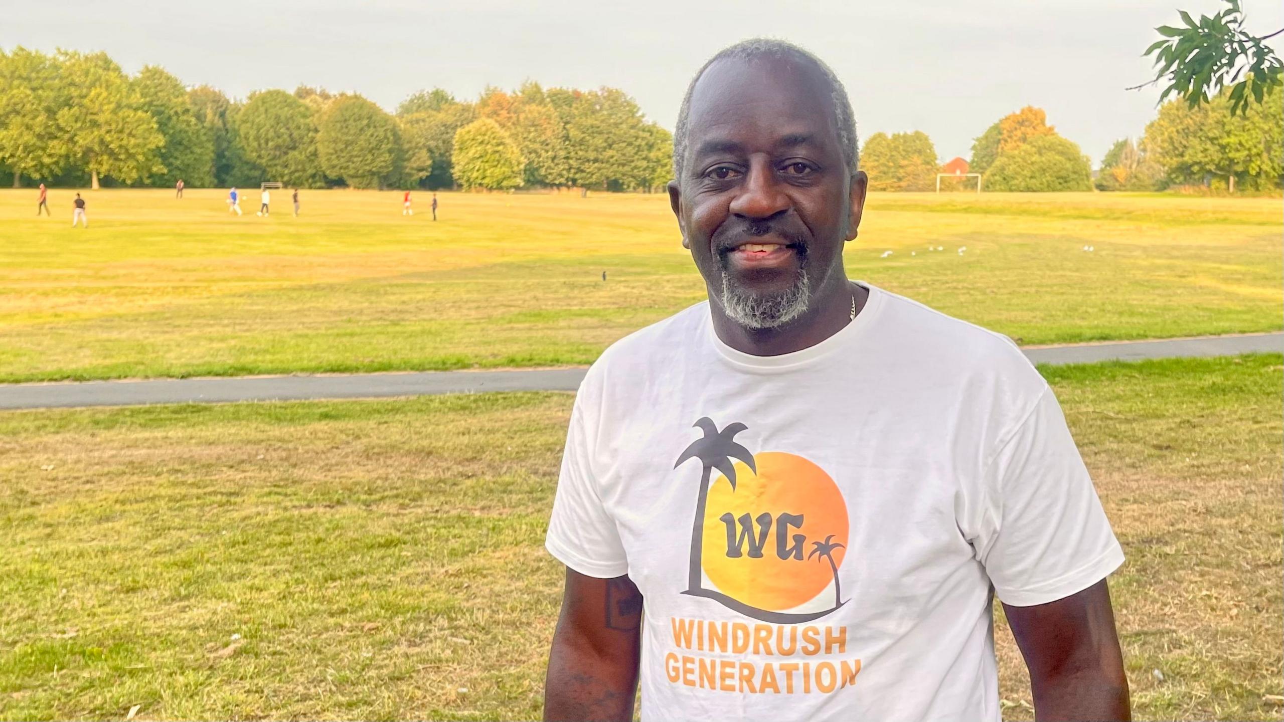A man with short grey hair and goatee beard stands in a park. His white tshirt has a sun, palm tree and the letters WG forming a logo. Beneath the logo it says WINDRUSH GENERATION. There are trees and people playing cricket in the background on a sunny day.