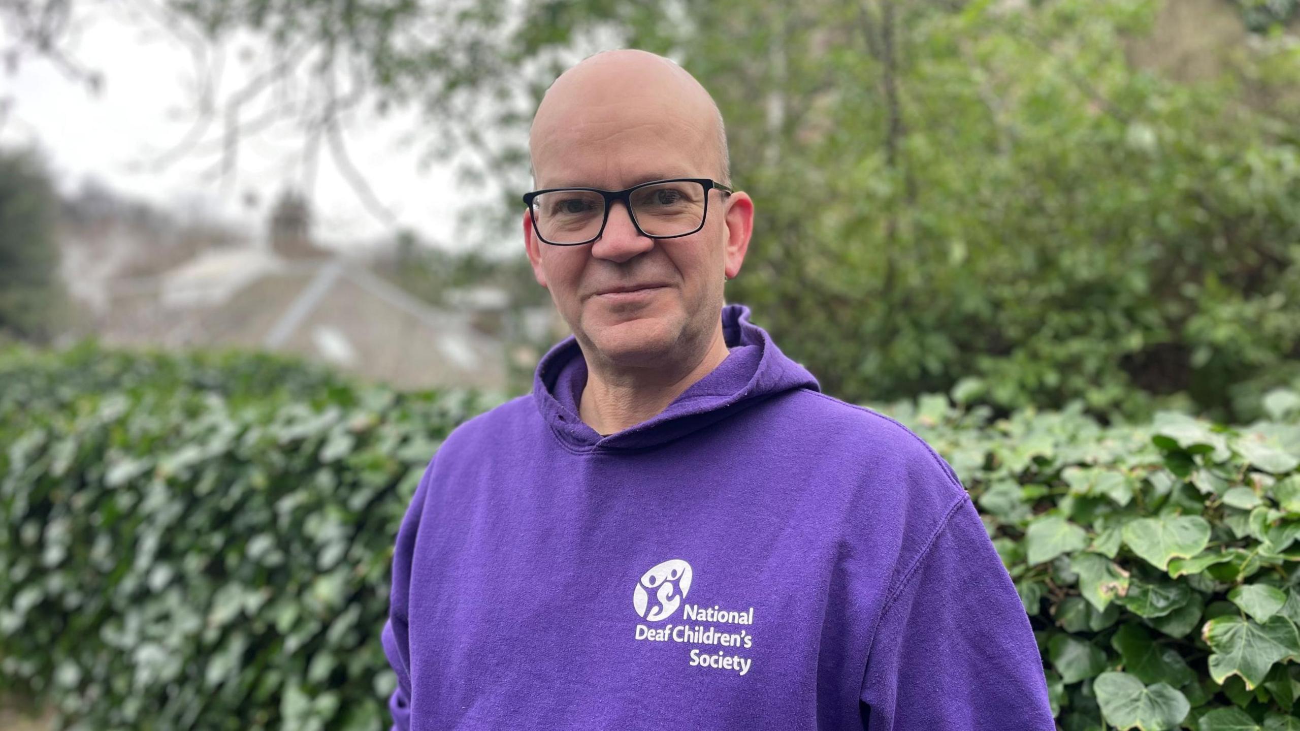 Mark Ballard wears a purple hoodie bearing the white logo of the National Deaf Children's Society, and is standing outside with a lush green hedge behind him and trees and houses in the distance. He wears dark-framed glasses