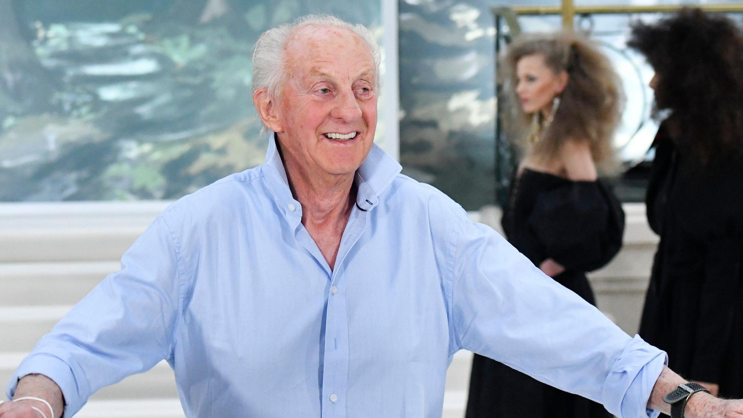 Paul Costelloe is is smiling broadly and has receding grey hair. He is wearing a blue open-neck shirt and his arms are stretched out on either side. behind him are three female models dressed in black.