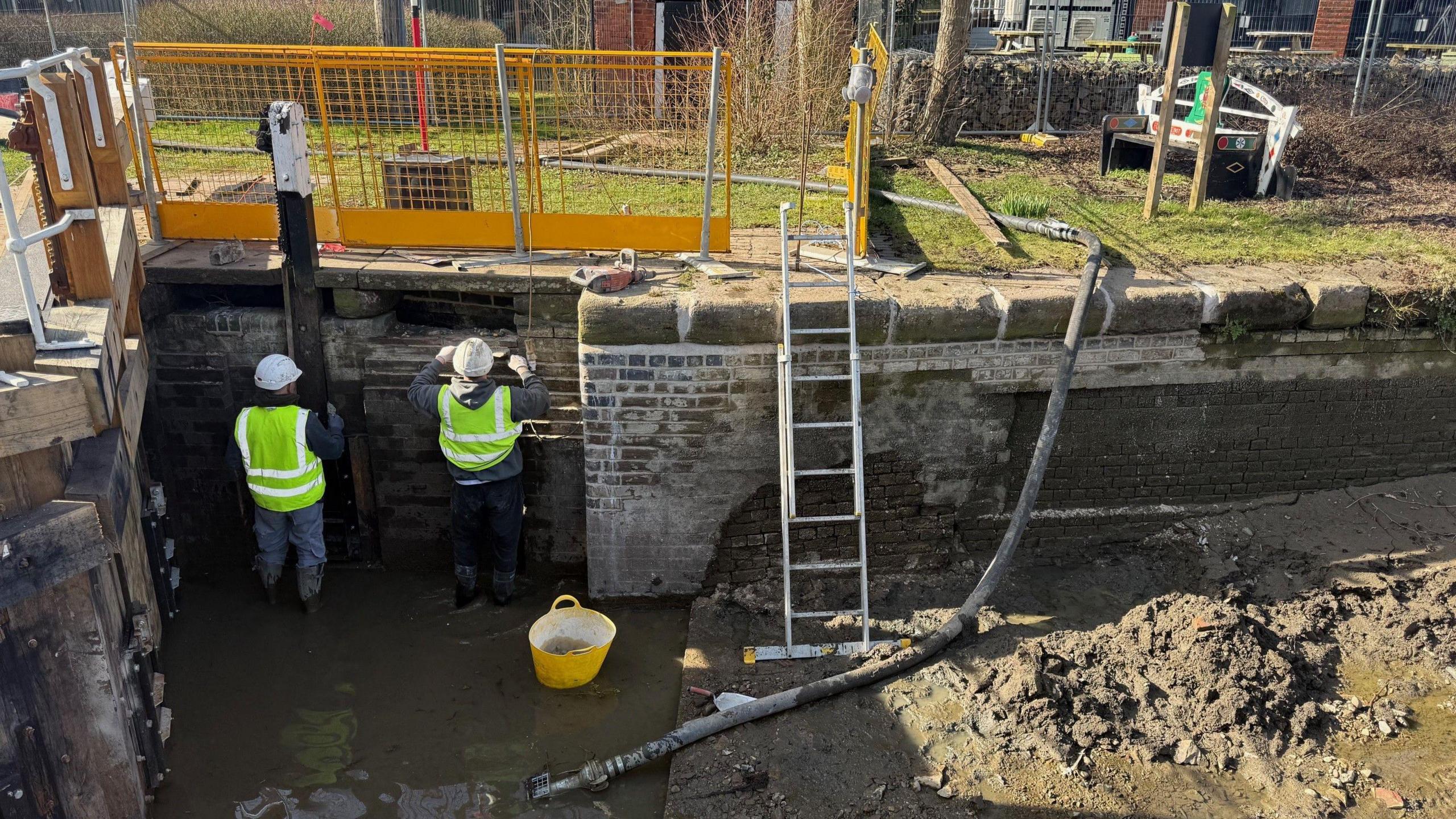 Two workers in yellow hi-vis working on the wall of a lock. There is a ladder propped up to the right, and a black pipe going from the towpath to the canal.  There are yellow fences on the towpath.