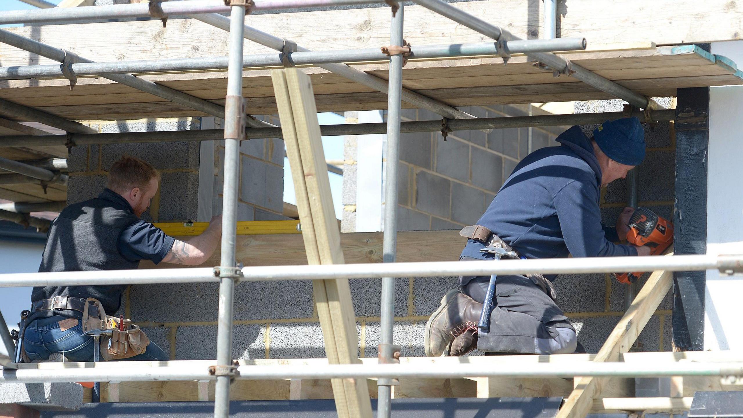 Two builders are working on a breeze block construction. They are kneeling on a scaffolded platform working on window apertures, one measuring with a spirit level and the other drilling.