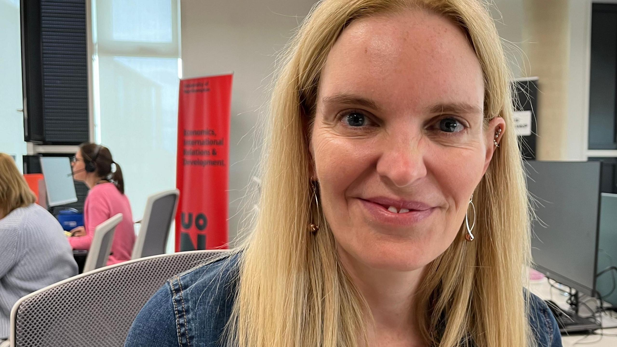 Anne-Marie Kilday, who has long, blonde hair and is wearing a blue denim top, sitting in an office chair and smiling at the camera. Desktop computers can be seen behind her and on the left of the picture two members of staff are taking calls.