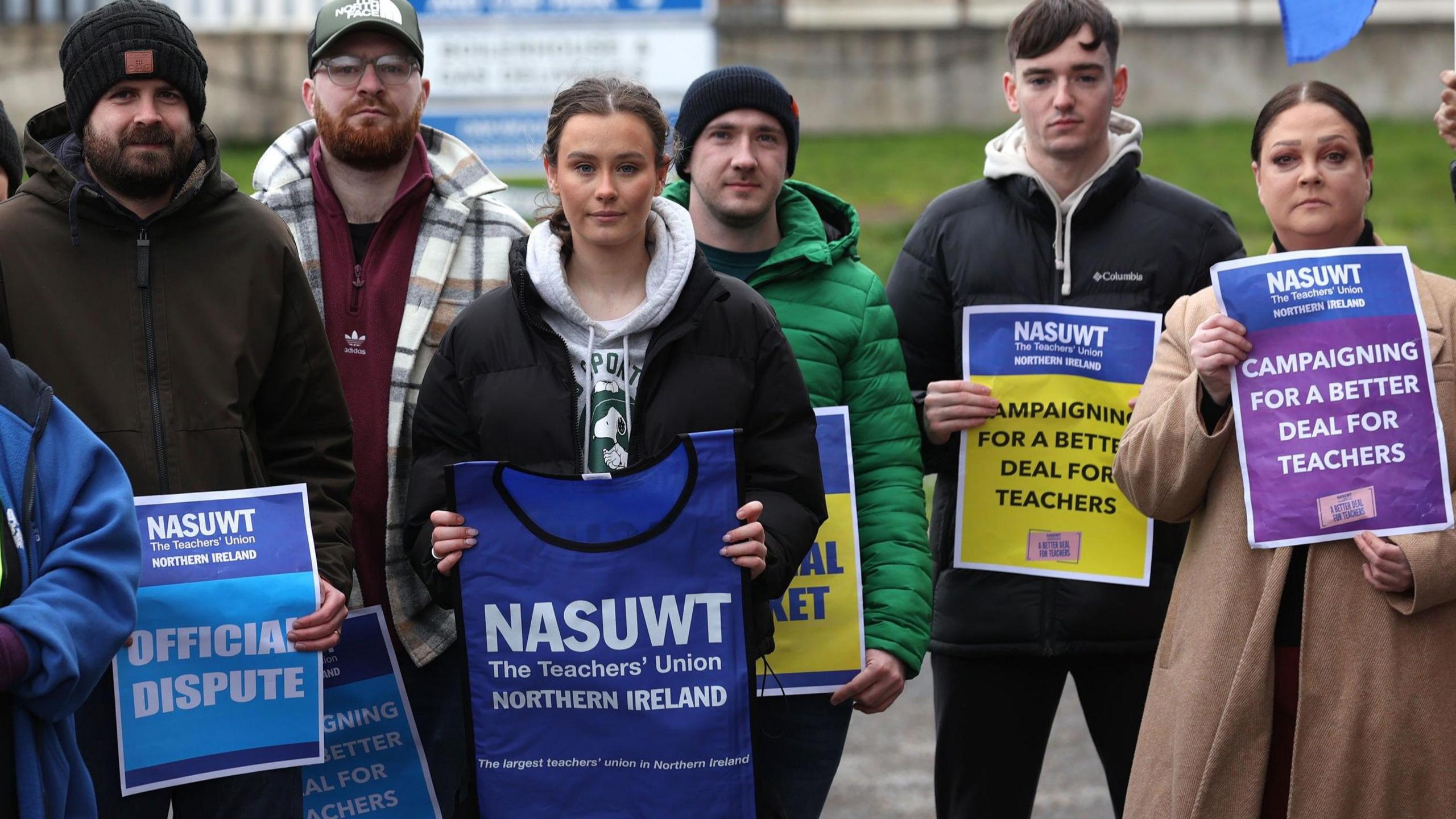 Six people stand on the picket line, holding up signs campaigning for a better deal for teachers. There are two girls and four boys. One girl holds a blue vest with white writing that says NASUWT, The Teacher's Union Northern Ireland.