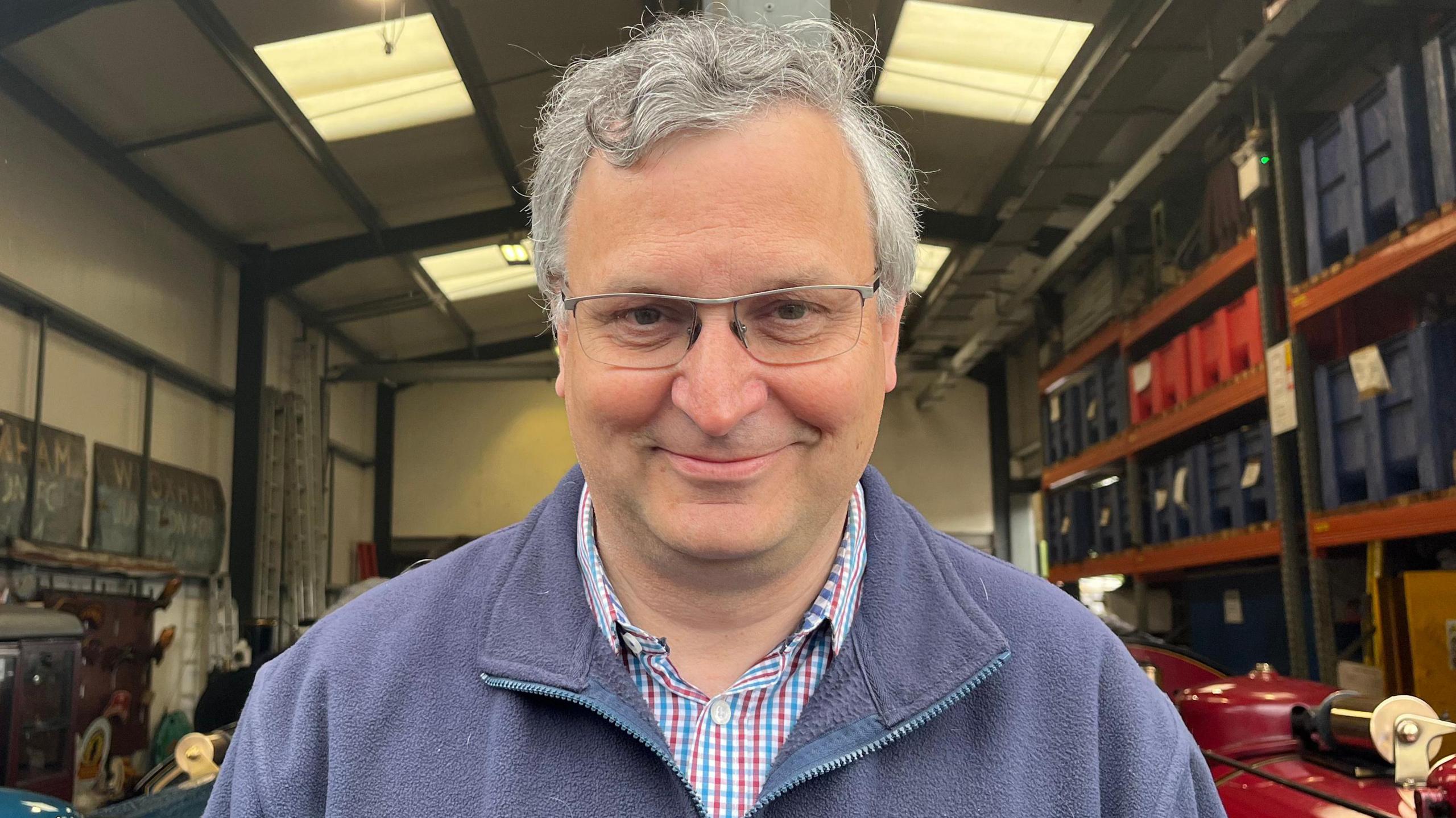 A man with grey hair looking directly at the camera and smiling. He is wearing a red and blue chequered shirt and a navy blue fleece.