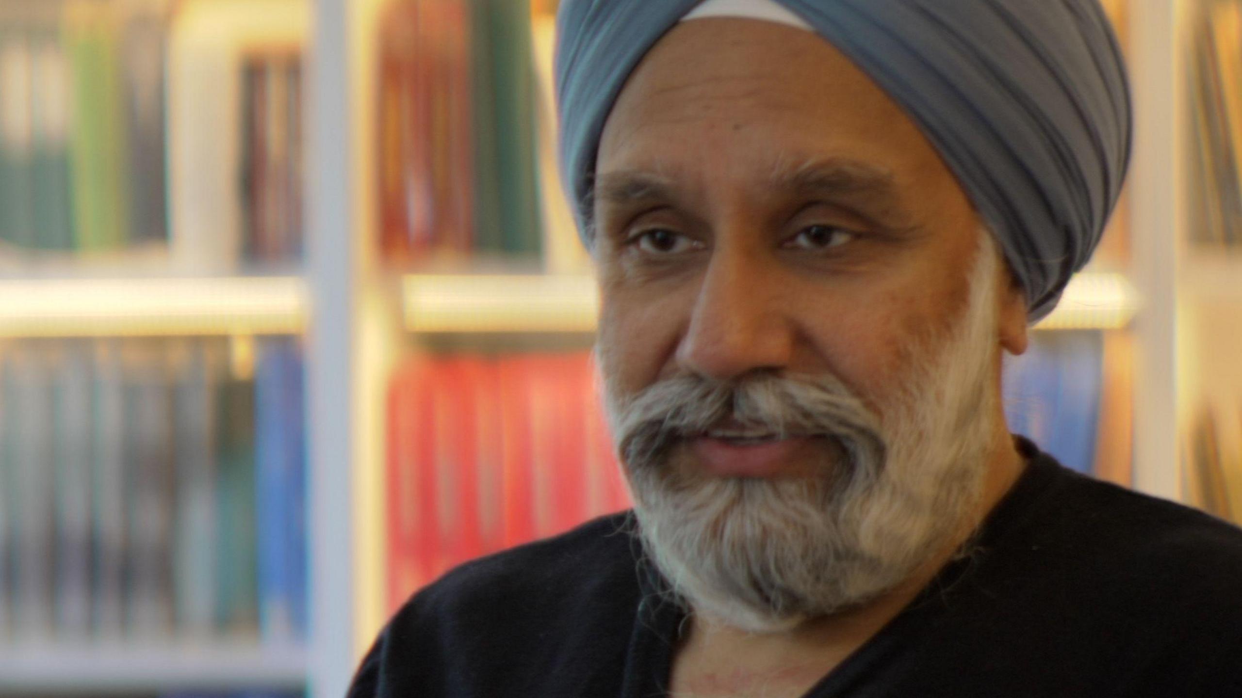 A middle aged Sikh man with a greying beard and a blue/grey turban stands in front of a bookcase. 