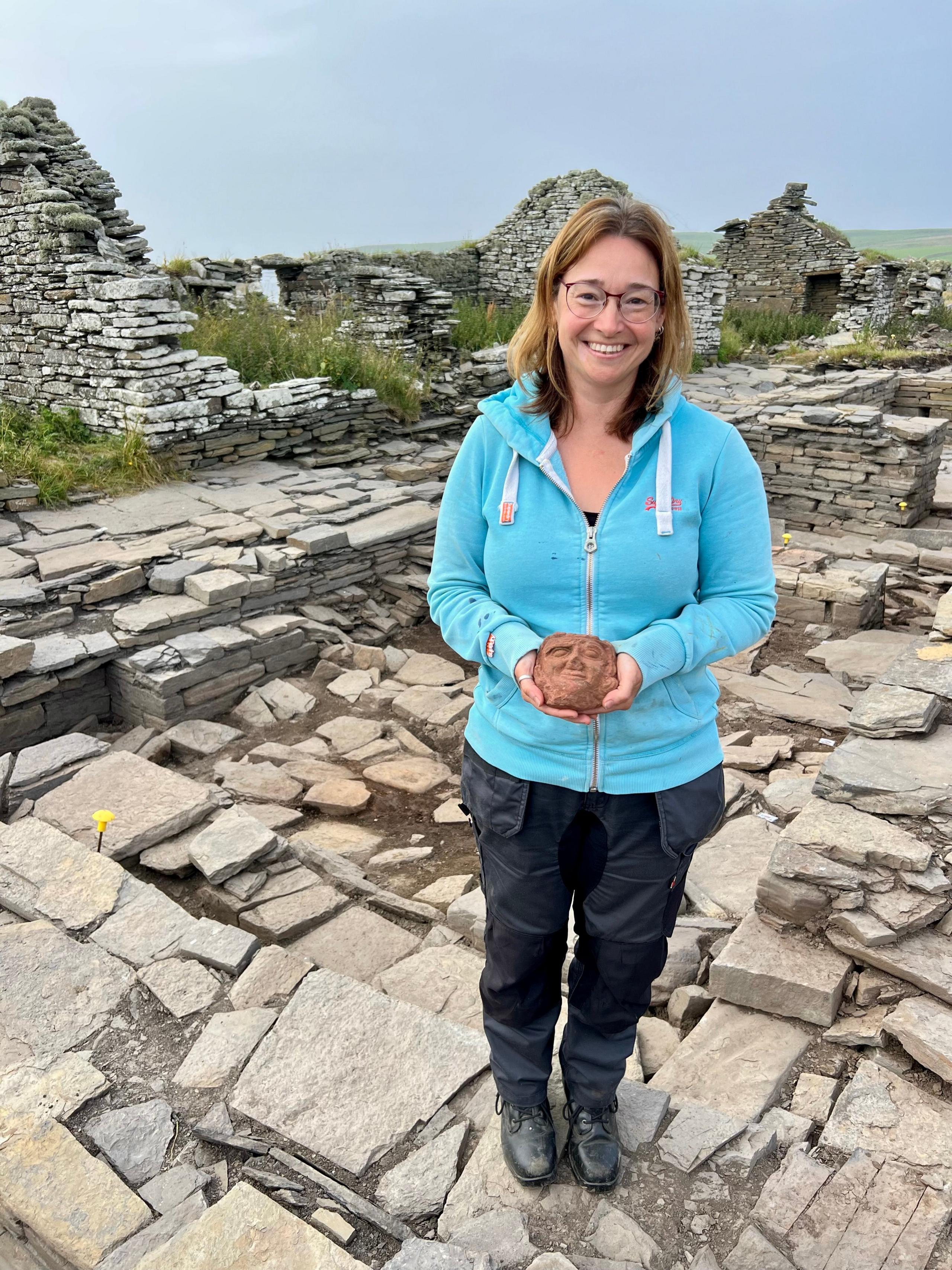 A woman with long brown hair and glasses in a light blue top with dark trousers sands in the middle of an archaeological dig site