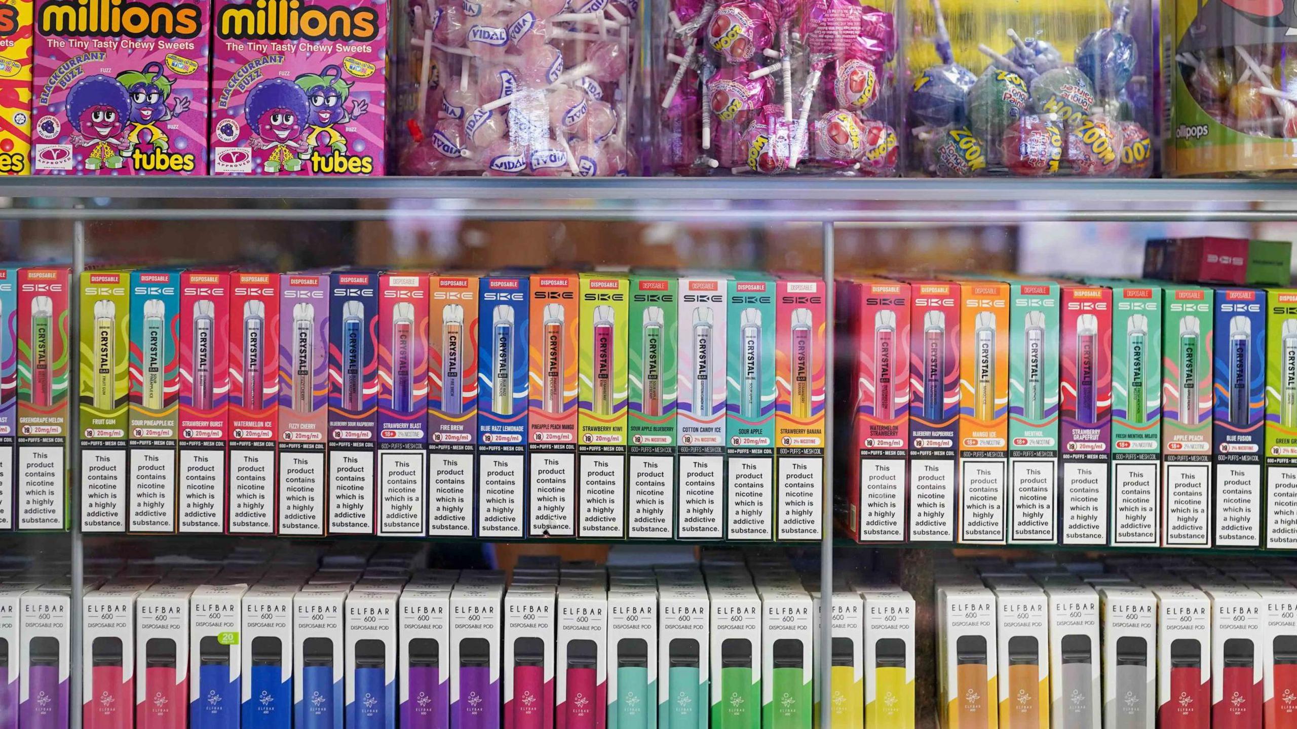 Two shelves of colourful single-use vapes in a shop. They are below sweets which have been placed on top of the glass cabinet they are in.