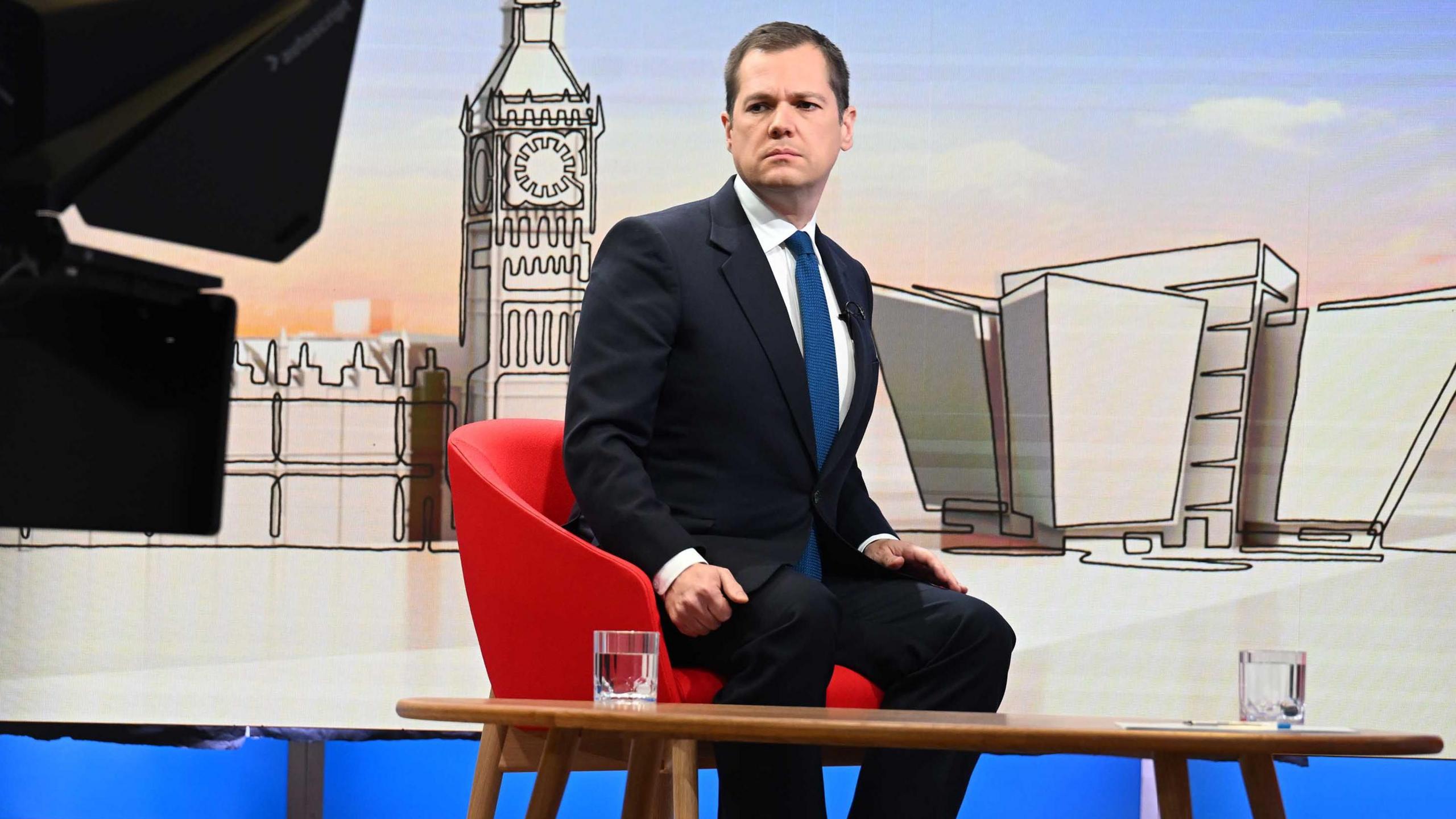 Robert Jenrick, who has short dark hair and is wearing a dark suit, white shirt and blue tie, sits in a BBC studio. There is a stylised image of the Palace of Westminster behind him and two glasses of water o a wooden table in front of him