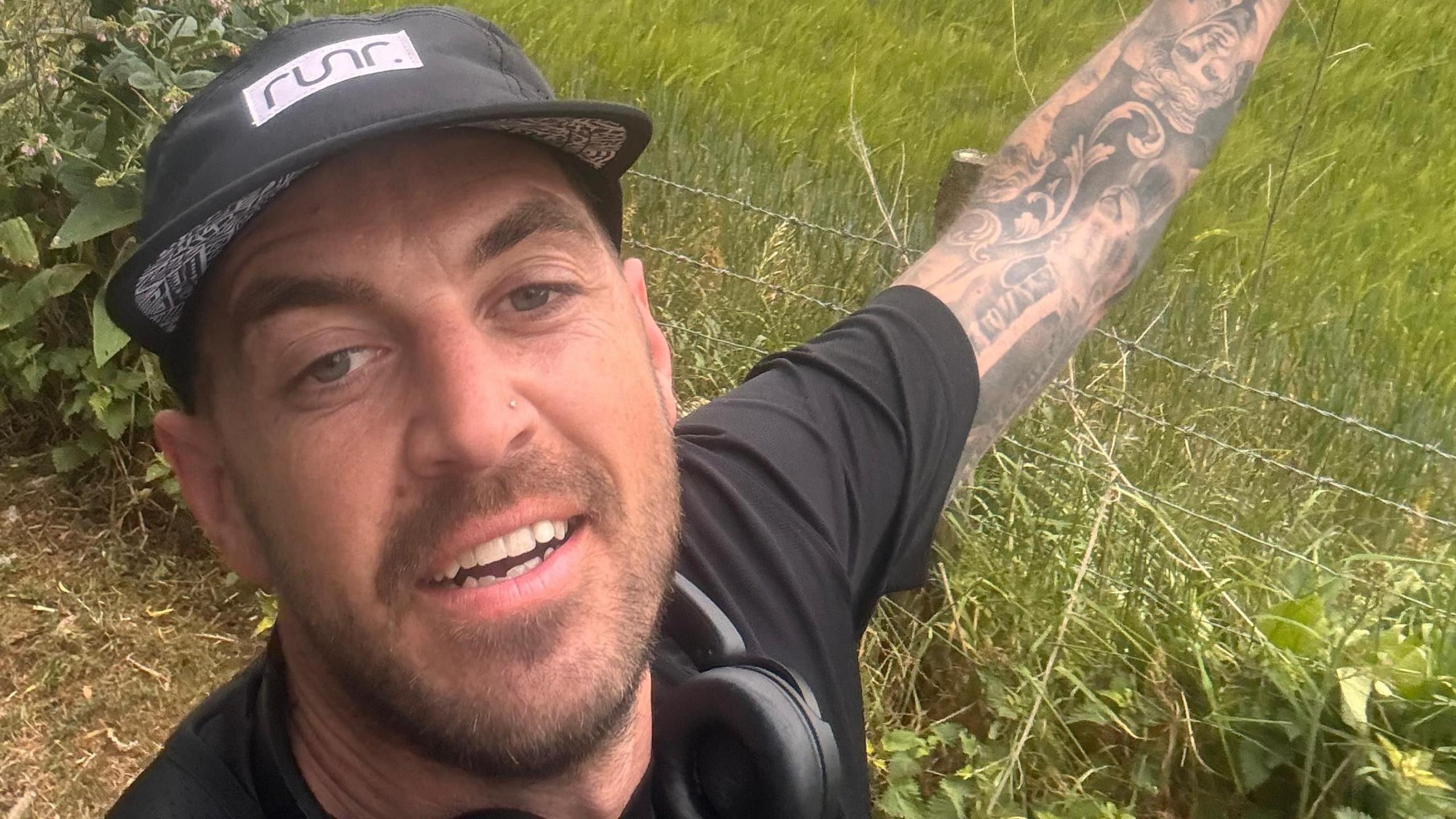 Joe Baker running in the countryside, holding his arm out, pointing to the green, open space. He is wearing a black T-shirt, a baseball cap and headphones around his neck.