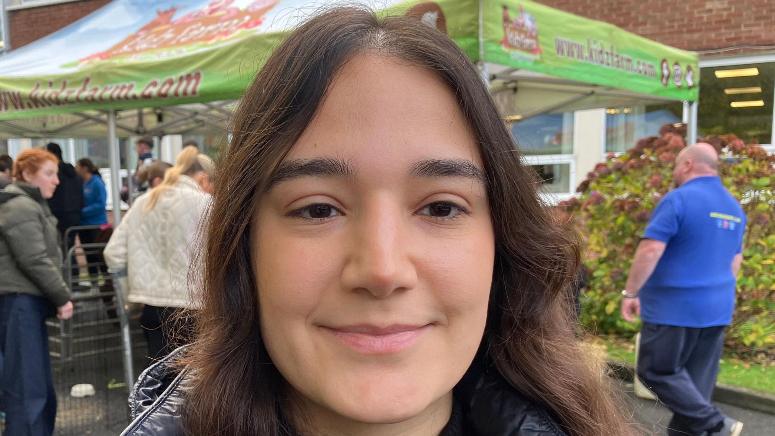 Lezin Baris has long brown hair and is wearing a black coat. She is outside in a courtyard with people behind her.