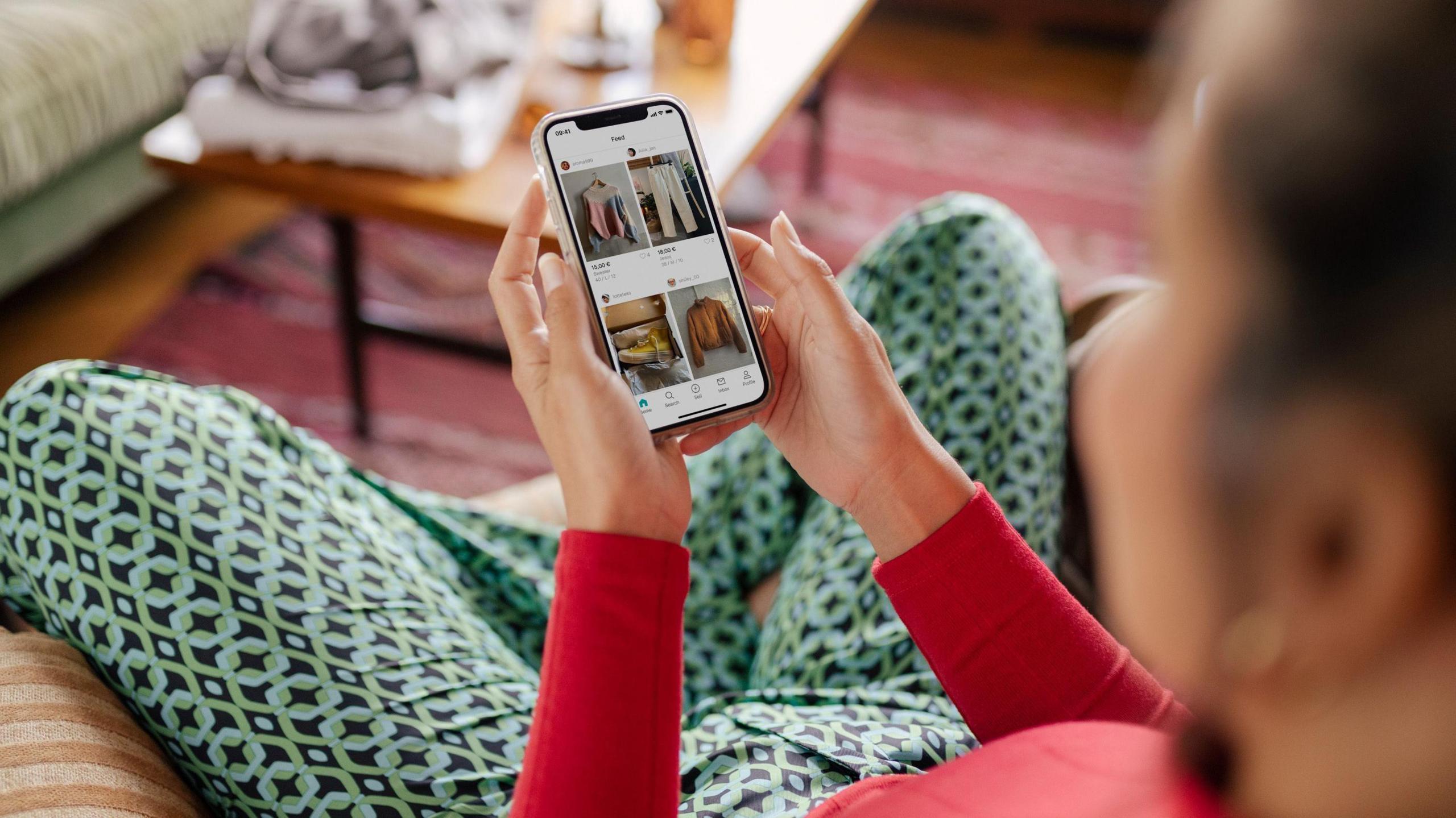 A young woman sits on an armchair holding a mobile phone she is using to browse items on Vinted 