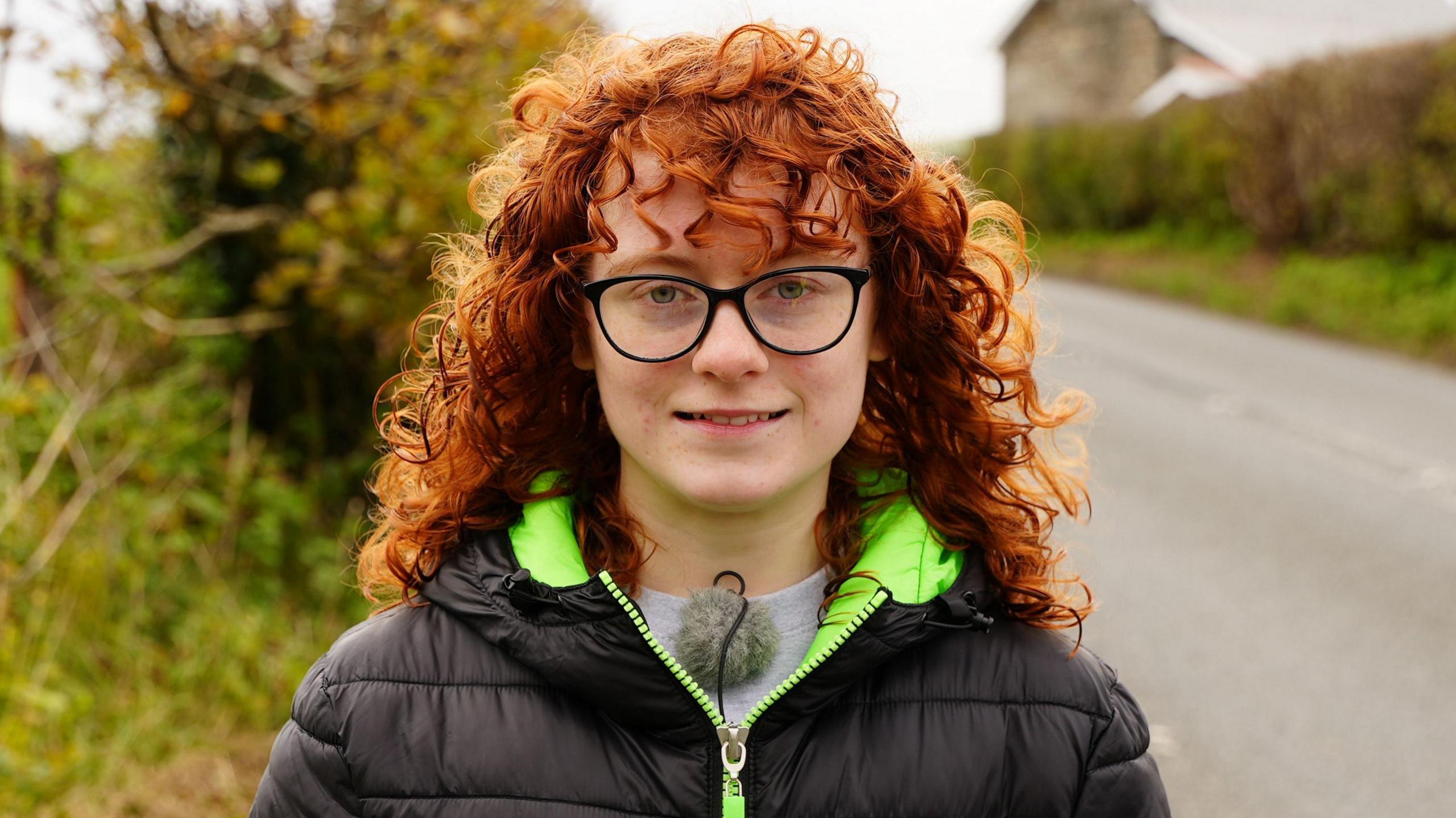 Merch gyda gwallt coch cyrliog a sbectol ddu yn sefyll o flaen gwrych gyda'r ffordd brysur y tu cefn iddi. 