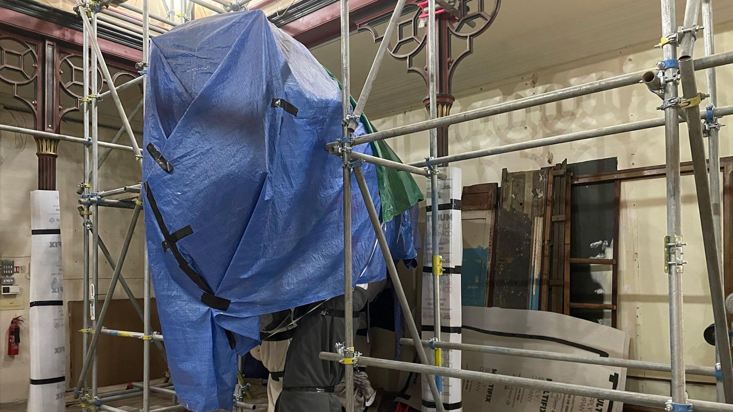 A photo of a mammoth statue that has remained inside the museum. It is wrapped in blue and green plastic sheets. Around it is scaffolding as well as other construction items.