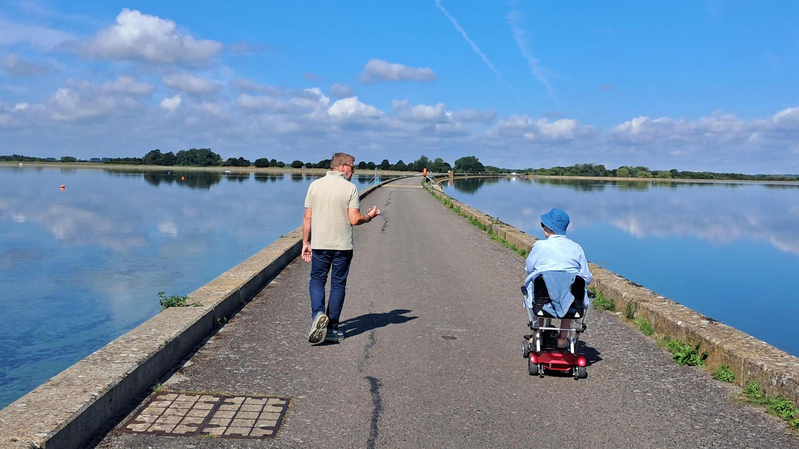 A man is walking on a tarmac path looking to his right talking to a person in a red mobility scooter. He is wearing a cream polo and jeans while the other person wears a blue top and bucket hat. The sun is shining and the sky is bright blue with a small number of white fluffy clouds. Either side of the tarmac path is water. The water is flat calm reflecting the sky. A line of trees can be seen on the horizon.