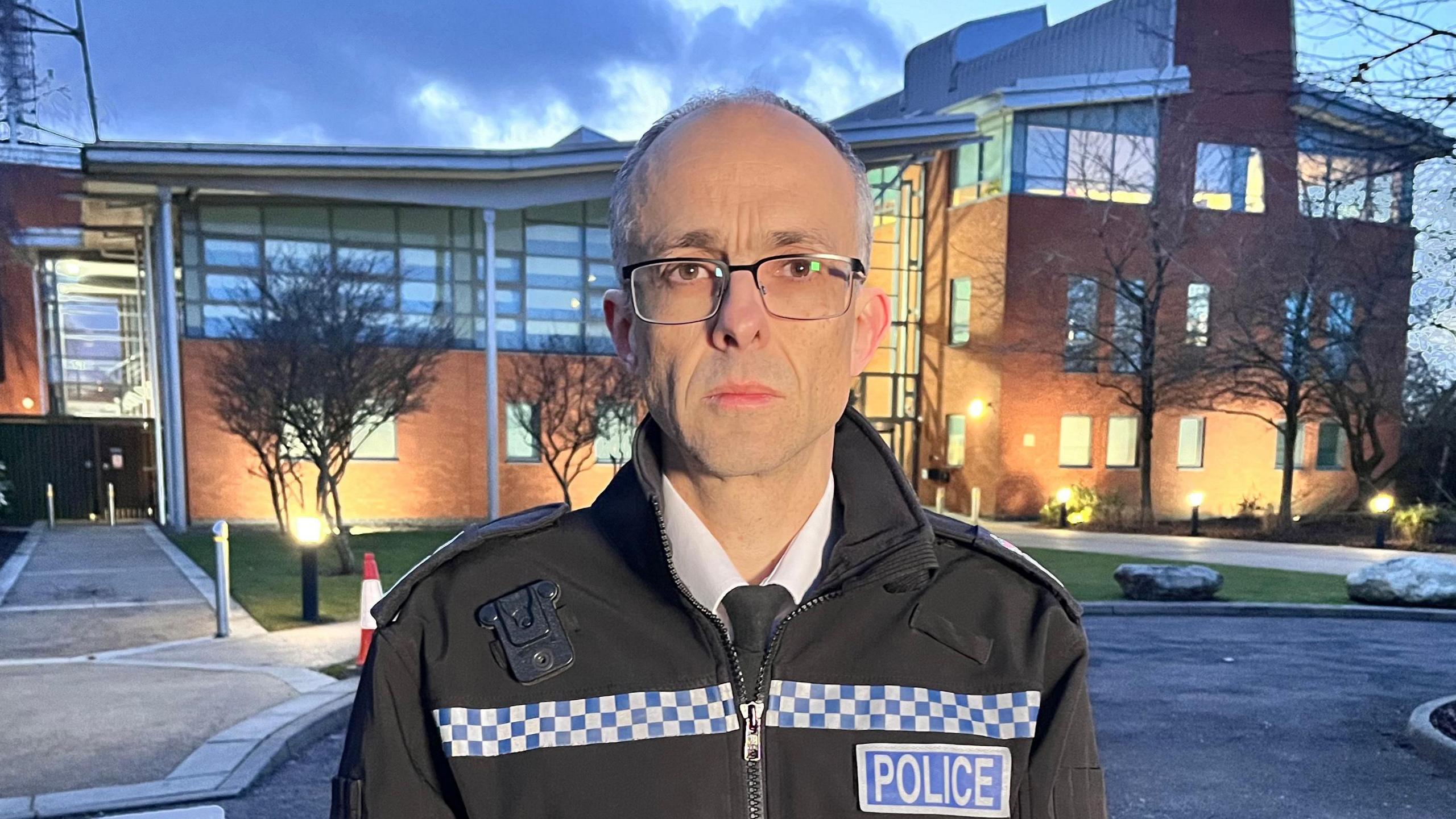 Paul Sanford is standing outside of the Norfolk Police Headquarters, a large, modern building. He's wearing a police jacket - labelled 'Police' - over a white shirt and black tie.