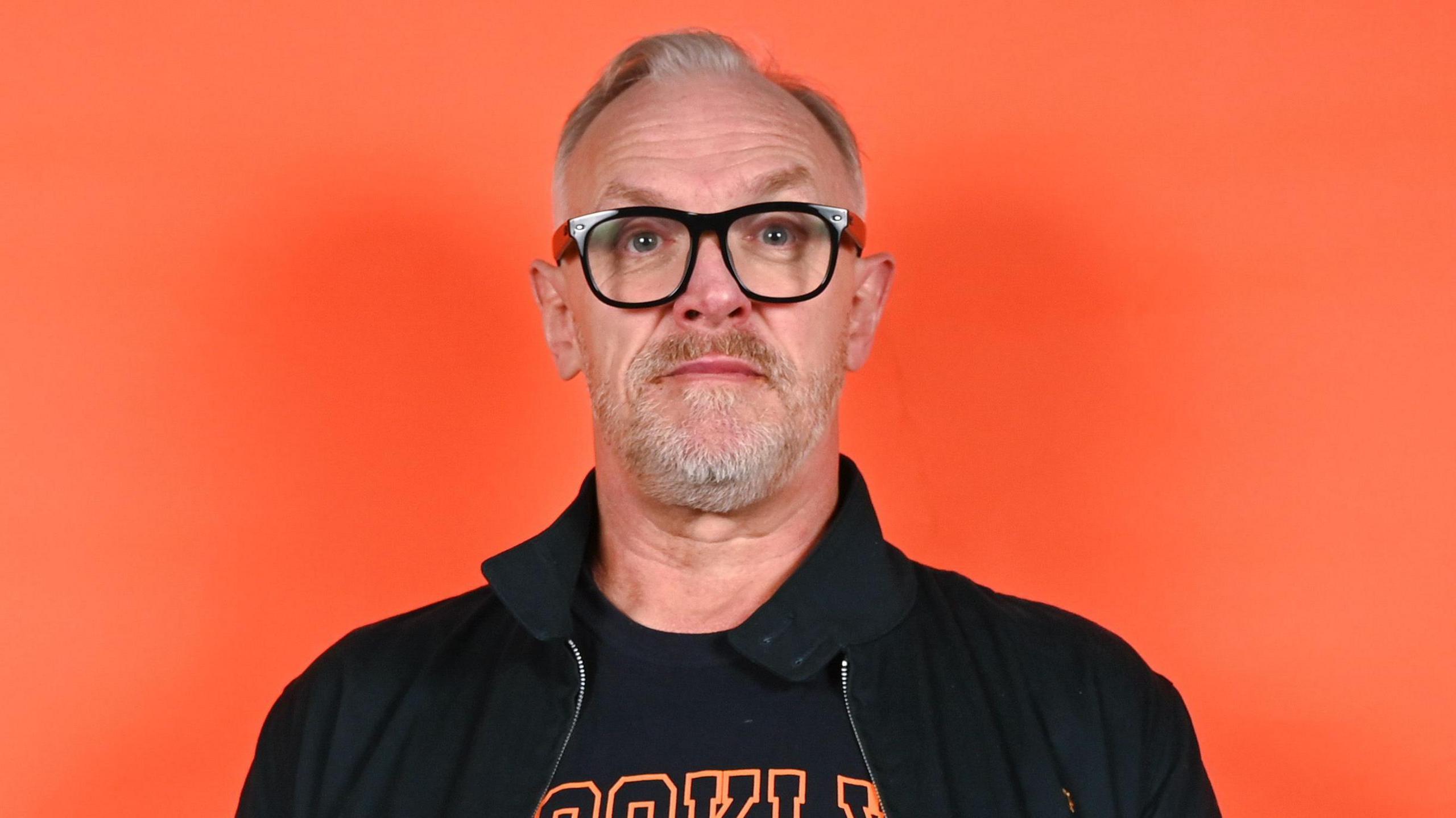 A man smiles at the camera while standing in front of an orange background. He has short grey hair, black glasses and is wearing a black jacket with a navy top on underneath.