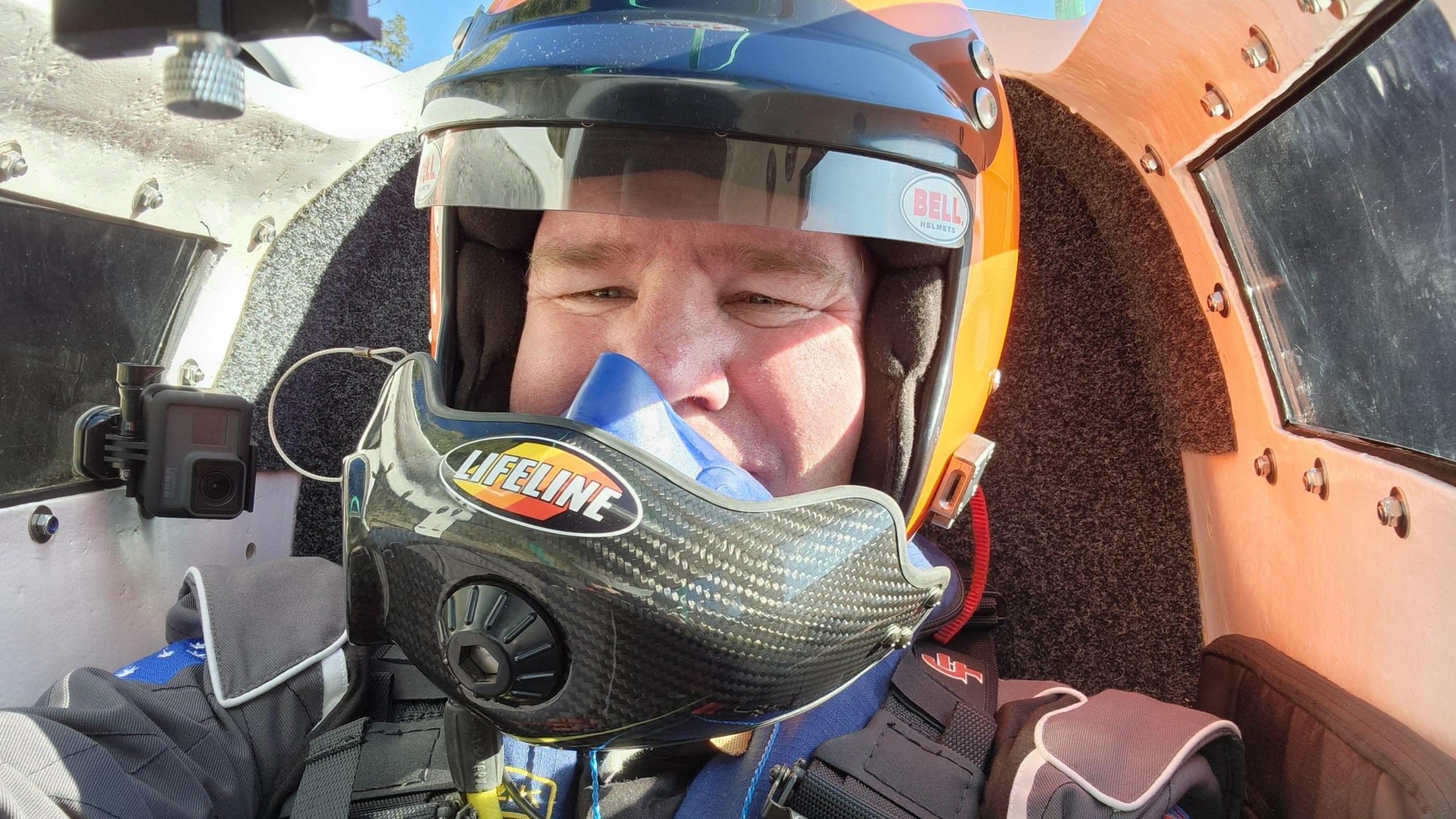 David Warby strapped into the cockpit of a hydroplane. He is wearing an orange crash helmet with a small black visor and black mouthpiece.