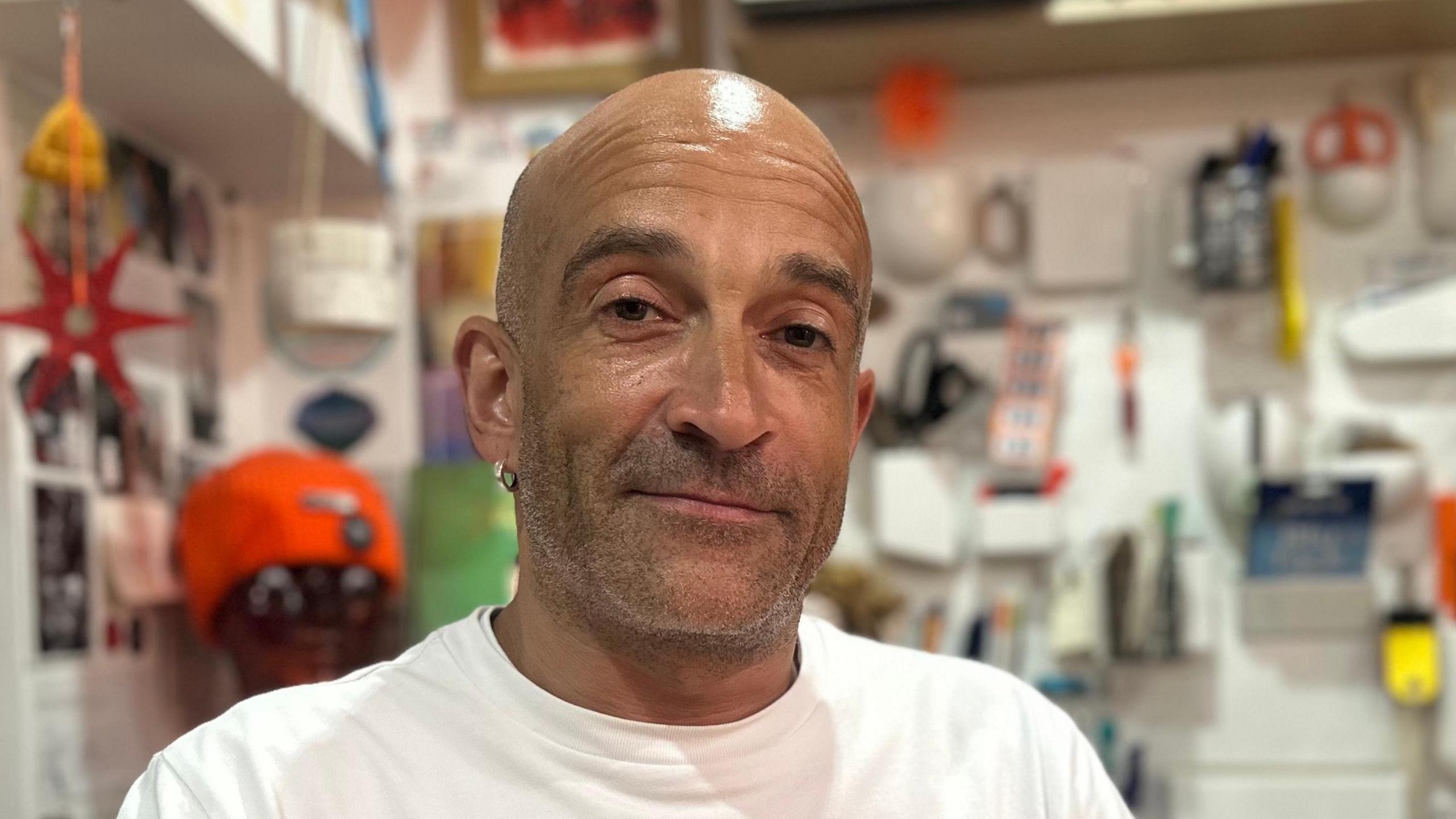 Andrew Marsh smiles at the camera in his book shop. He is bald and has a shaved beard. He wears a white T-shirt. The background is blurred.