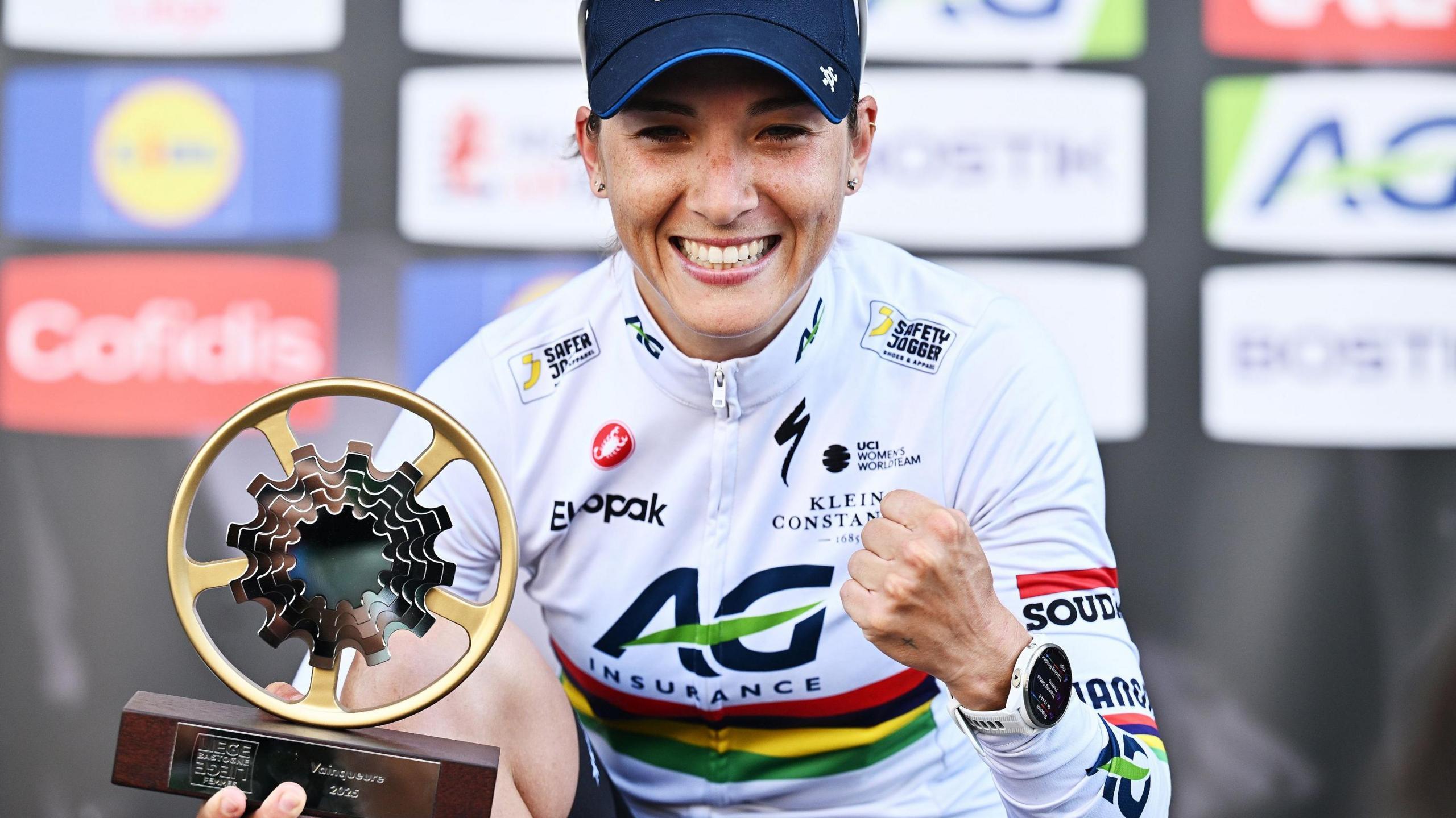 Kim Le Court wears a blue cap and cycling jersey as she smiles and clenches her left fist in celebration while using her other hand to hold the Liege-Bastogne-Liege cycling race trophy which is a golden wheel with spokes leading into a darkly-coloured derailleur gear mechanism