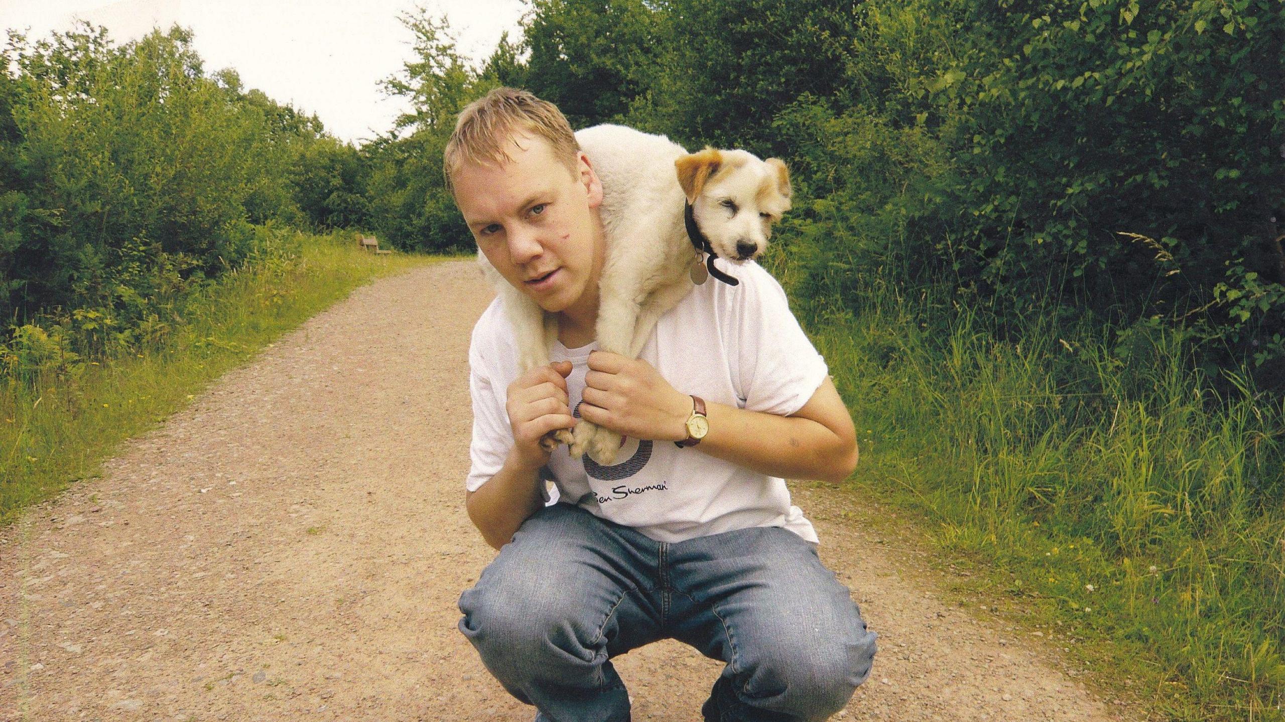 A man with blonde hair, wearing light blue jeans and a white t-shirt, squats over a path. He has a small white dog with light brown ears around his neck, and he is holding the dog's legs. The dog wears a black collar with a silver tag. The man and the dog are on a path, which is surrounded on both sides by grass and large green bushes.