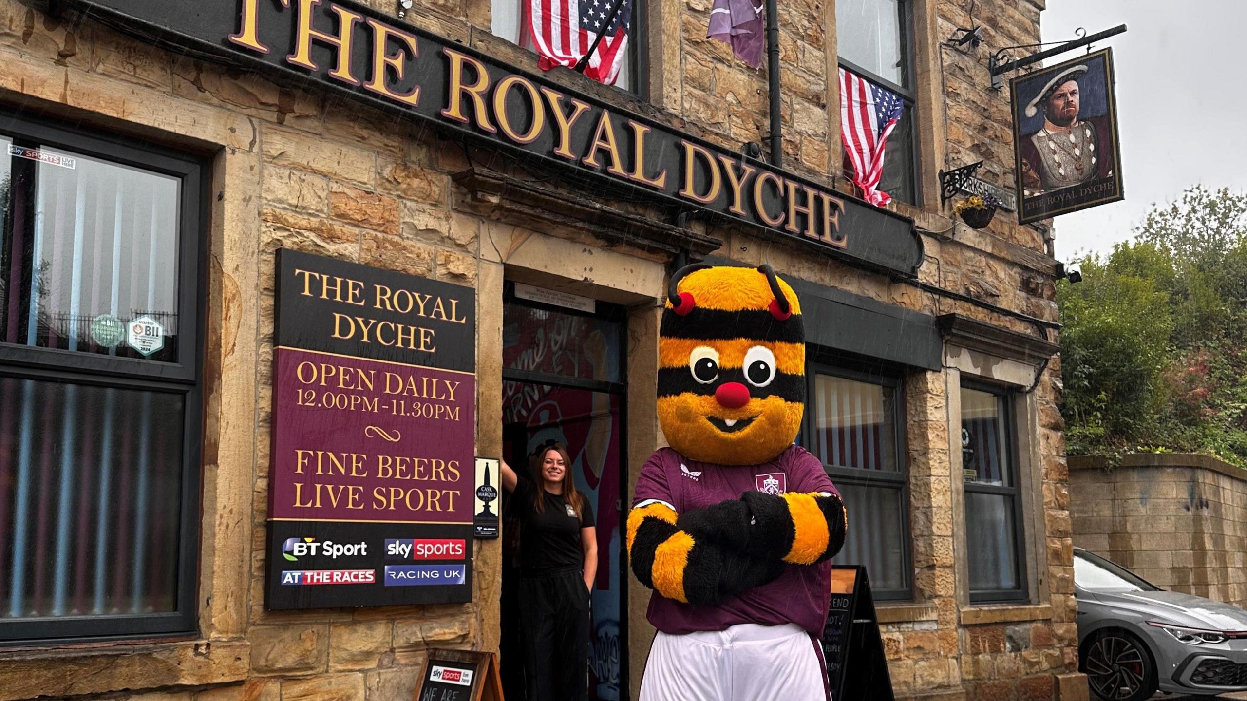 Burnley FC mascot Bertie Bee standing outside Burnley pub The Royal Dyche with landlady Justine Bedford dressed in black in the doorway smiling.
