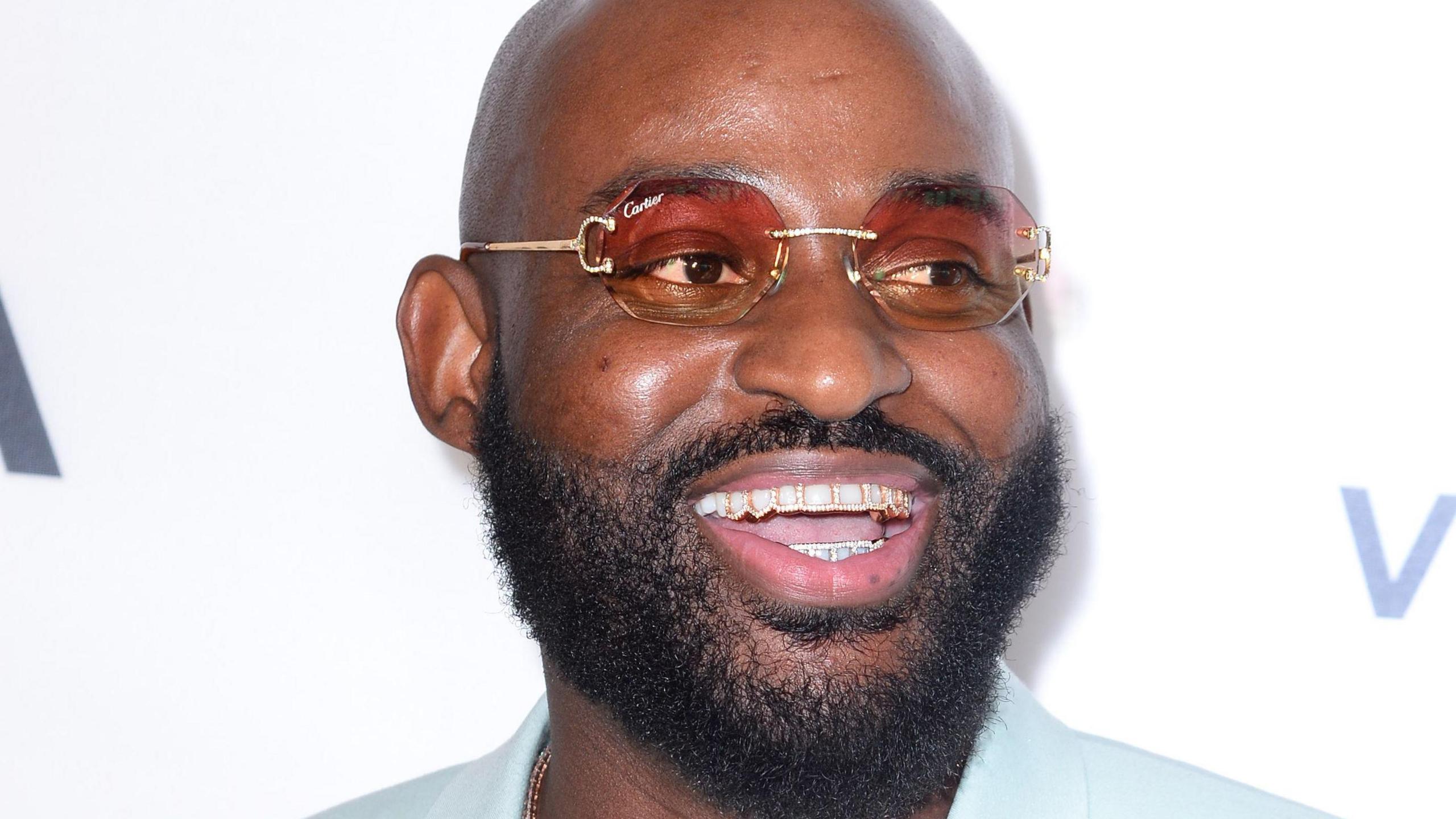 Posty at the 15th GRM Gala. Posty is a black man with a shaved head and short beard. He wears a pale blue shirt and rimless designer sunglasses and smiles to reveal embellished grills on his teeth 