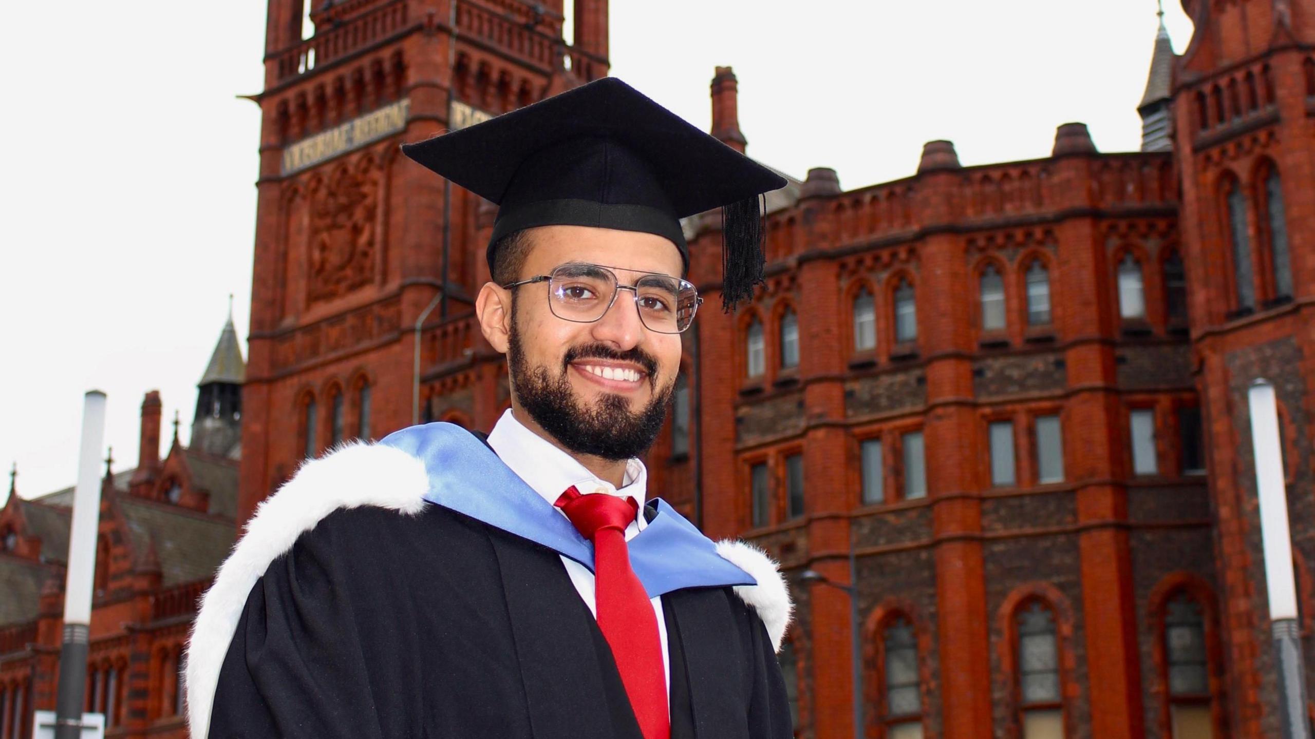 Hamza Al-Huseini stands outside the University of Liverpool's Victoria Building in his graduation cap and gown. He wears glasses and has a short beard.