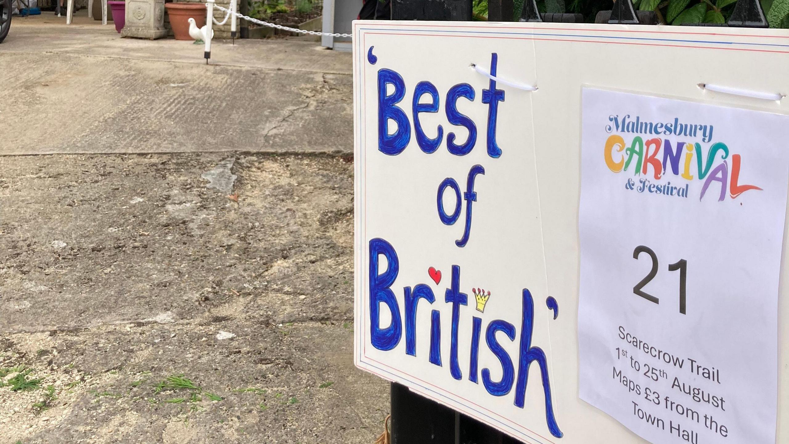 A sign reading: "Best of British". It is written in blue letters. There is a paper sign next to it advertising the Malmesbury Carnival and Scarecrow Festival.