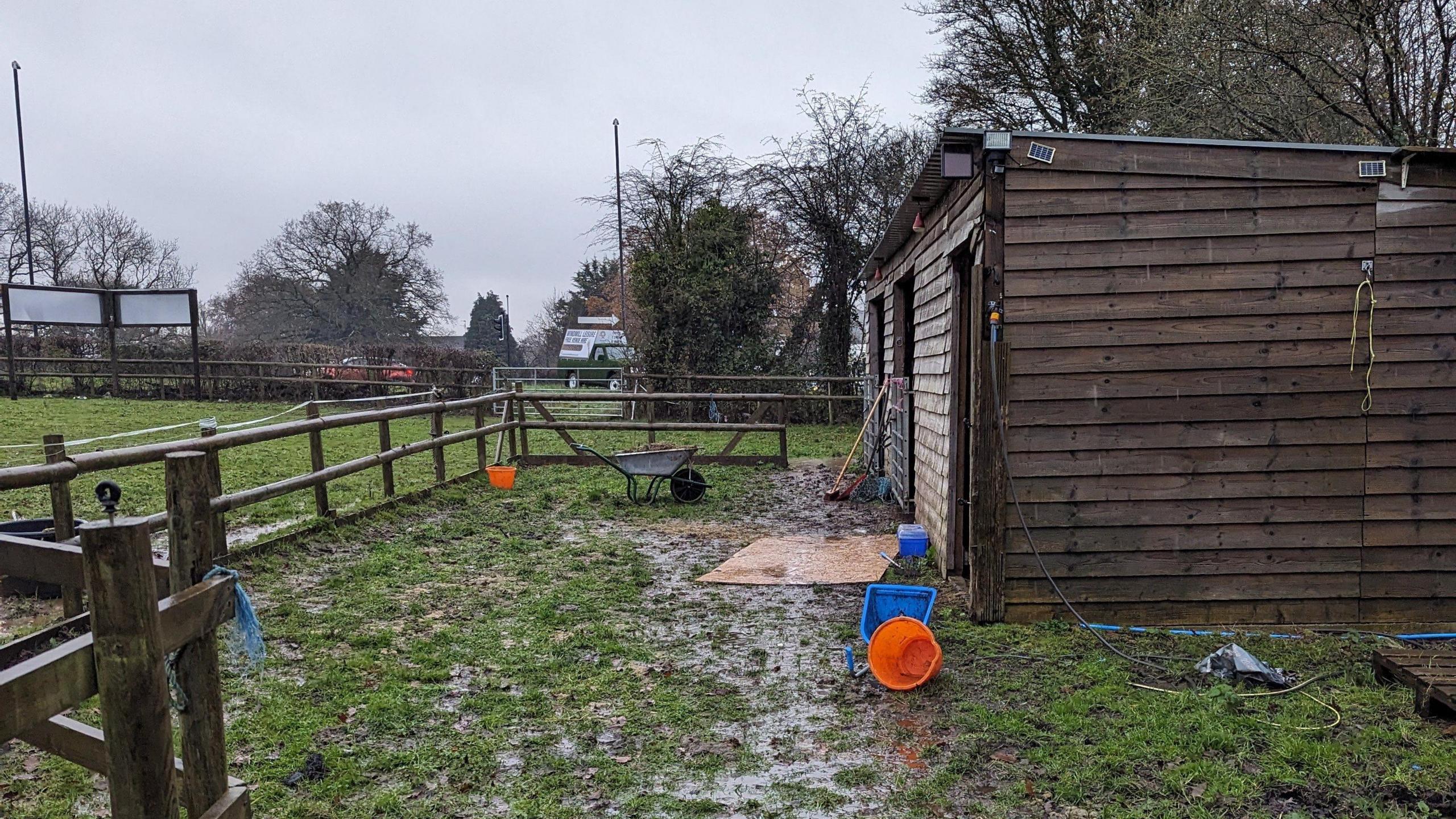 A muddy field in Westerleigh where Shane Paul Sims kept his two horses. A small stable building can be seen, with a wheelbarrow and other supplies nearby. There is a fence around the small area. 