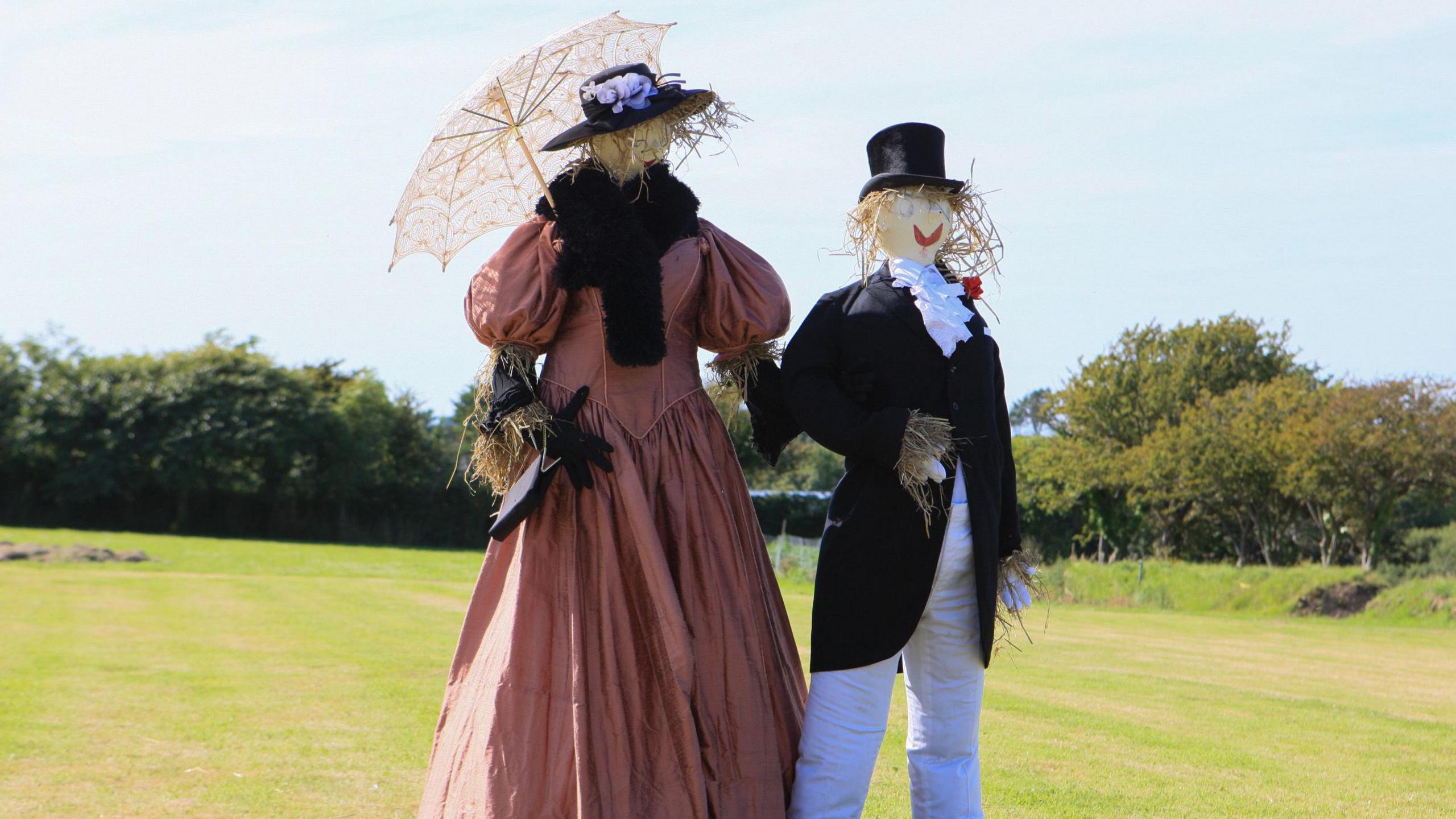 A picture of two scarecrows holding hands at a previous event. One is wearing a black and white suit and one is wearing a red dress.