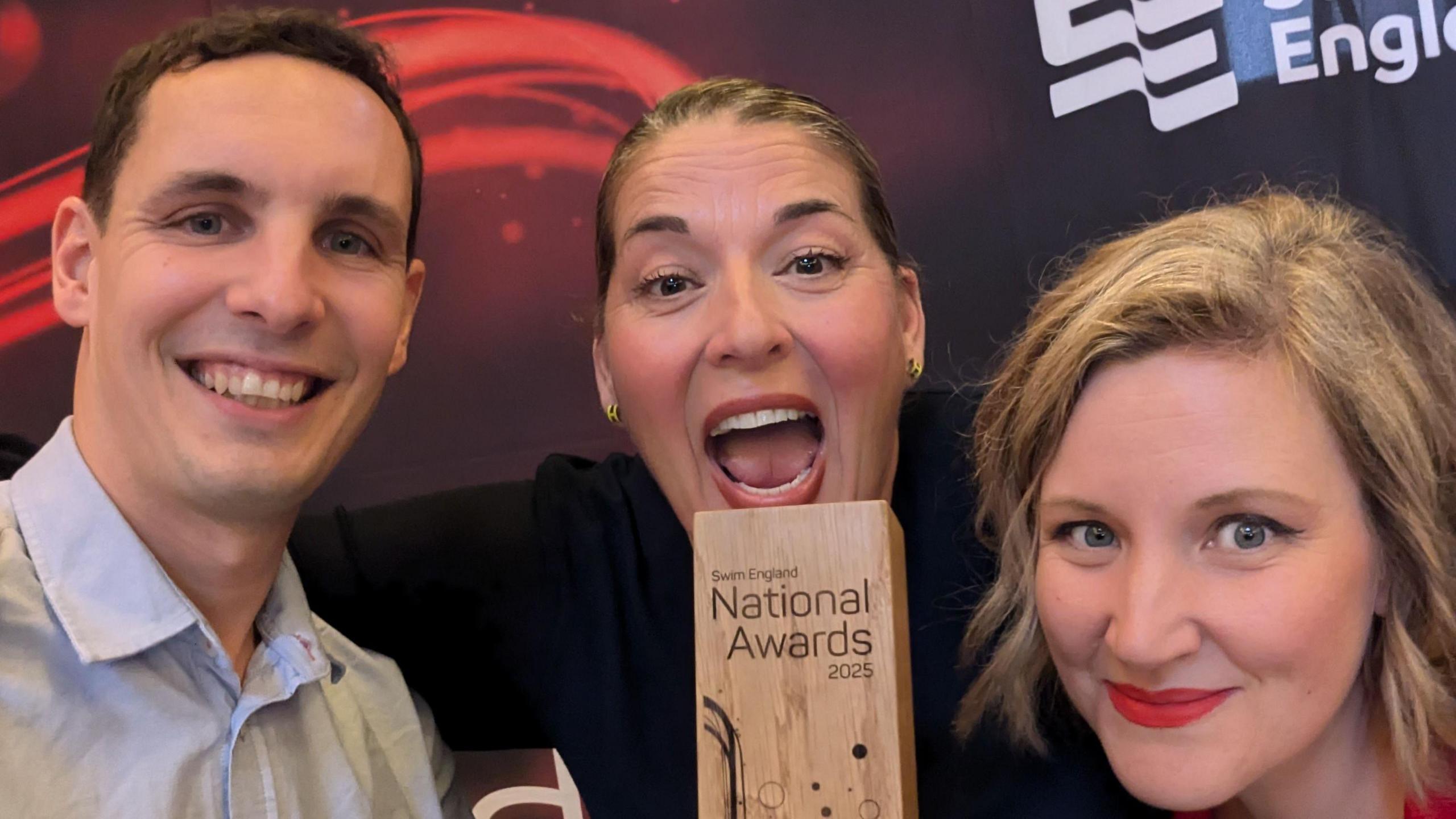 Three members of the swimming academy smiling with a wooden trophy between them