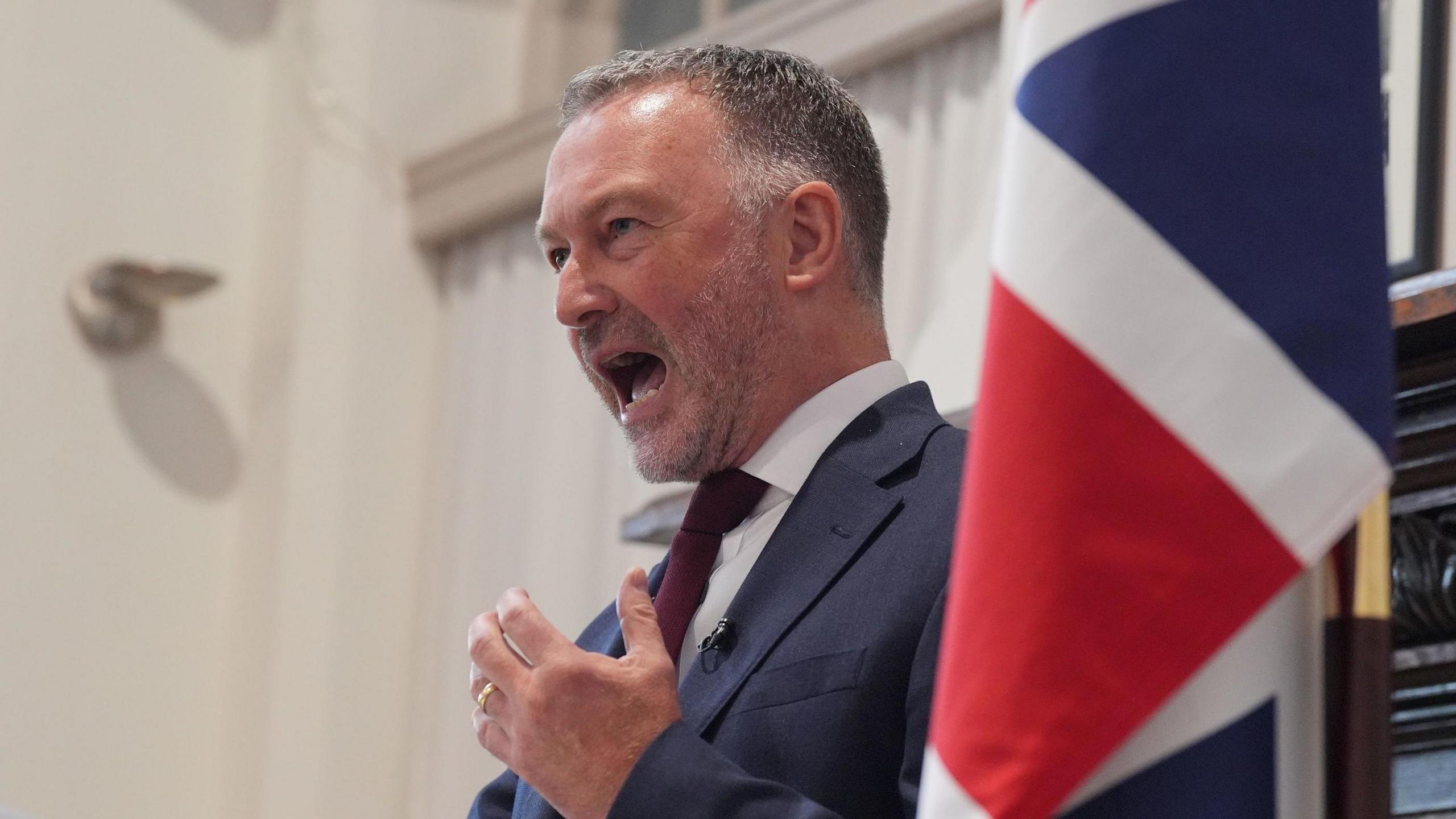 Steve Reed speaking at an event with his left hand raised towards his chest. He is wearing a blue suit, white shirt and burgundy tie and is standing next to a UK flag.