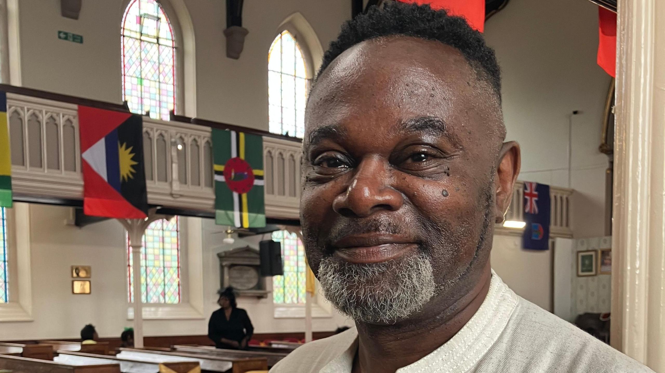 A man stands inside a church. Flags from around the country can be seen hanging in the background as well as rows of wooden pews.
