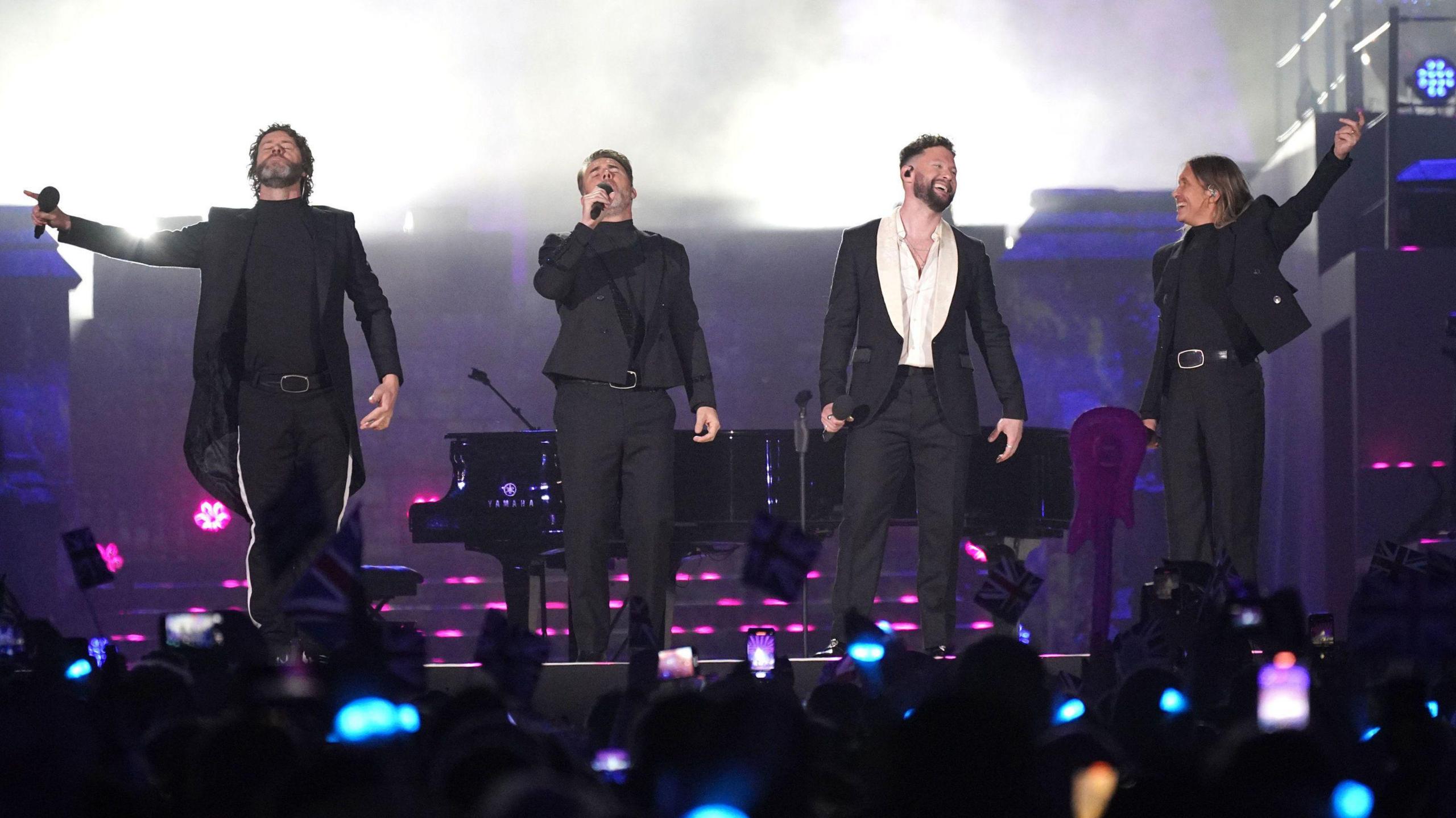Four male singers dressed in black suits standing in a row on stage in front of a live audience. A grand piano and lighting effects can be seen in the background.