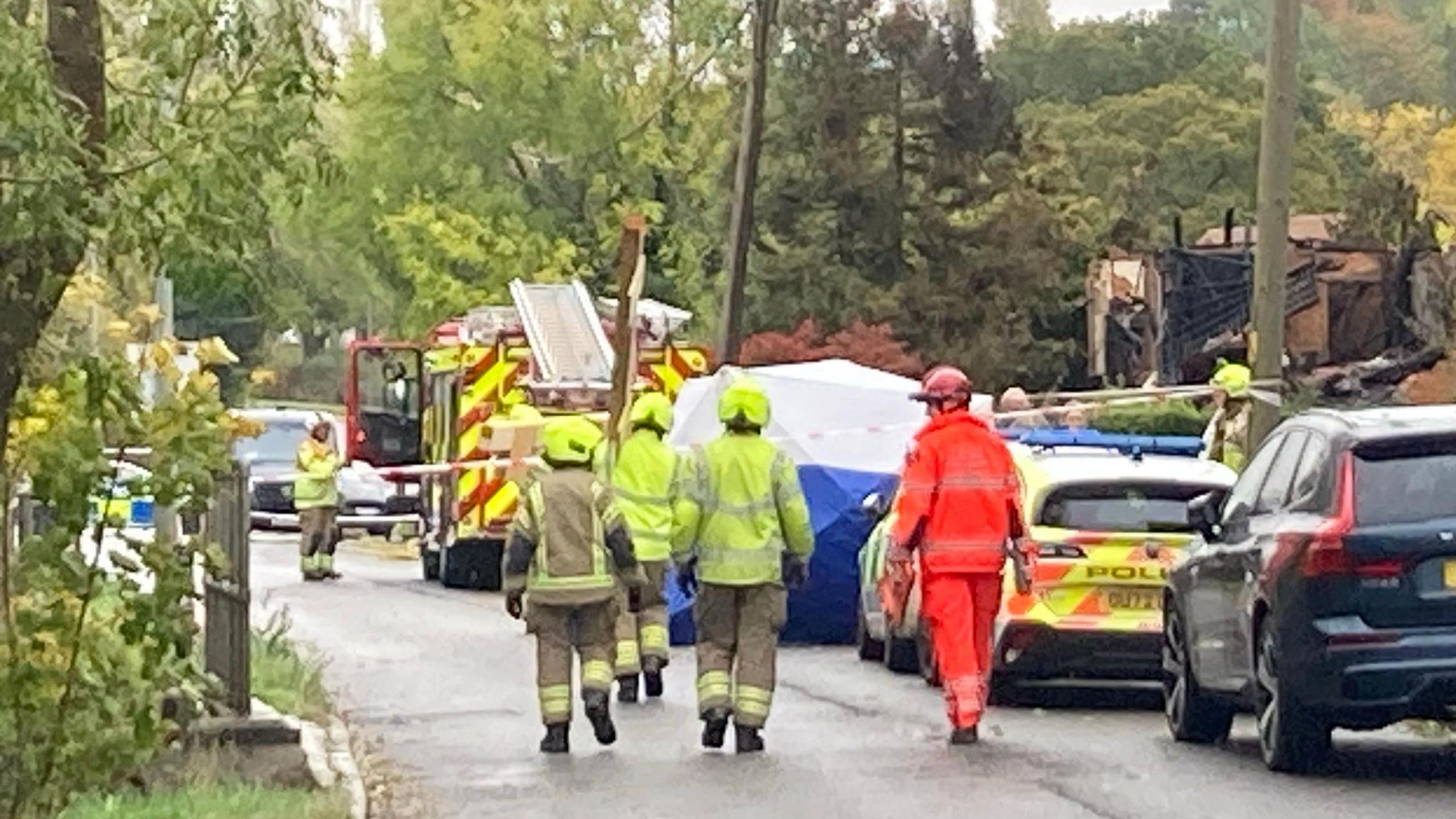 A number of emergency service workers in road in Bedford. They are walking away from the camera to a house that has been destroyed. A number of cars are  in the road, a police tent and a fire engine is in the street. The road has a number of trees and is in a residential area. 