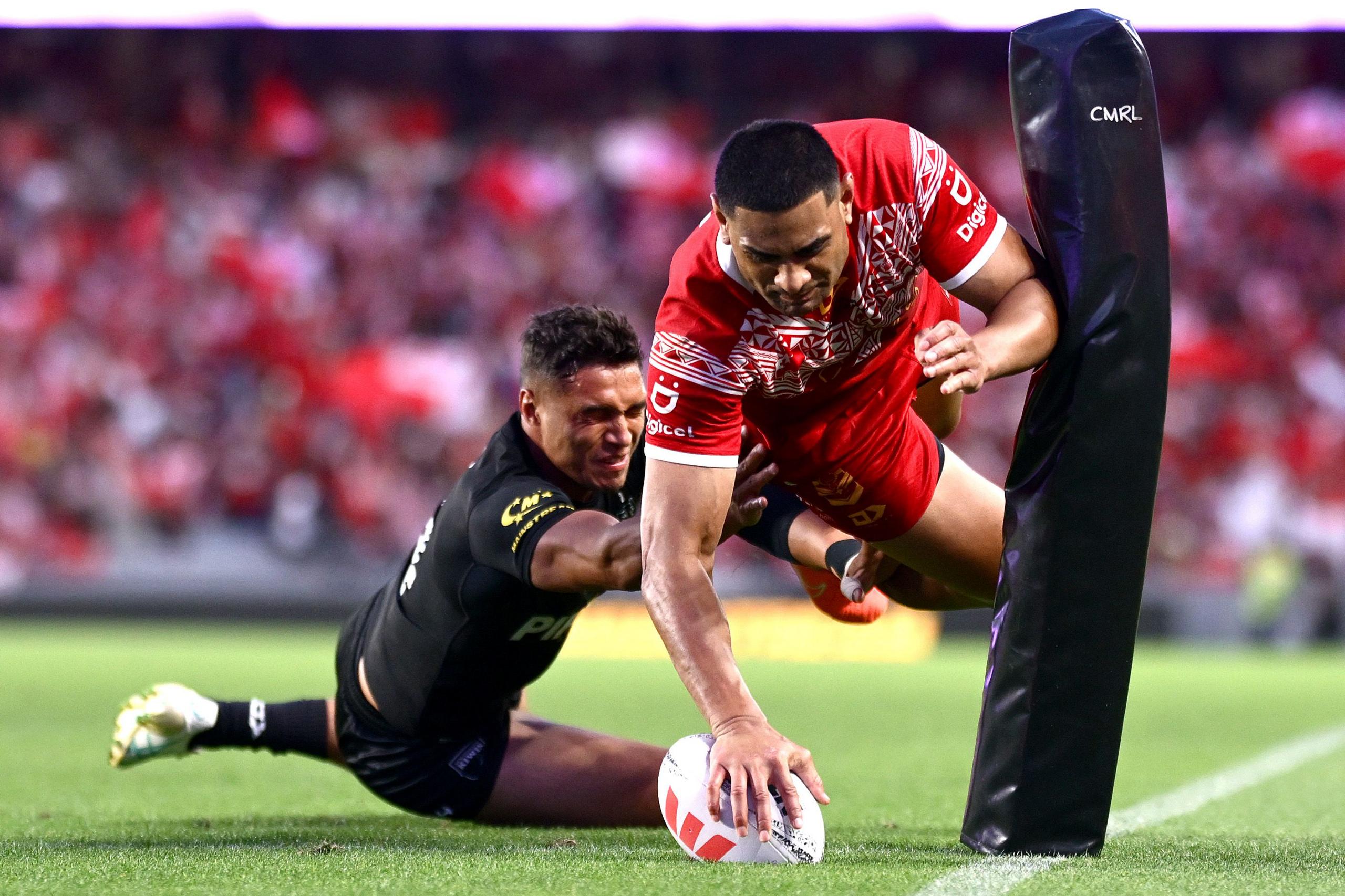 A player in a red jersey dives toward the try-line, extending an arm to ground the ball near the goalpost. An opposing player in a black uniform reaches out attempting a tackle. The stadium background is filled with spectators.
