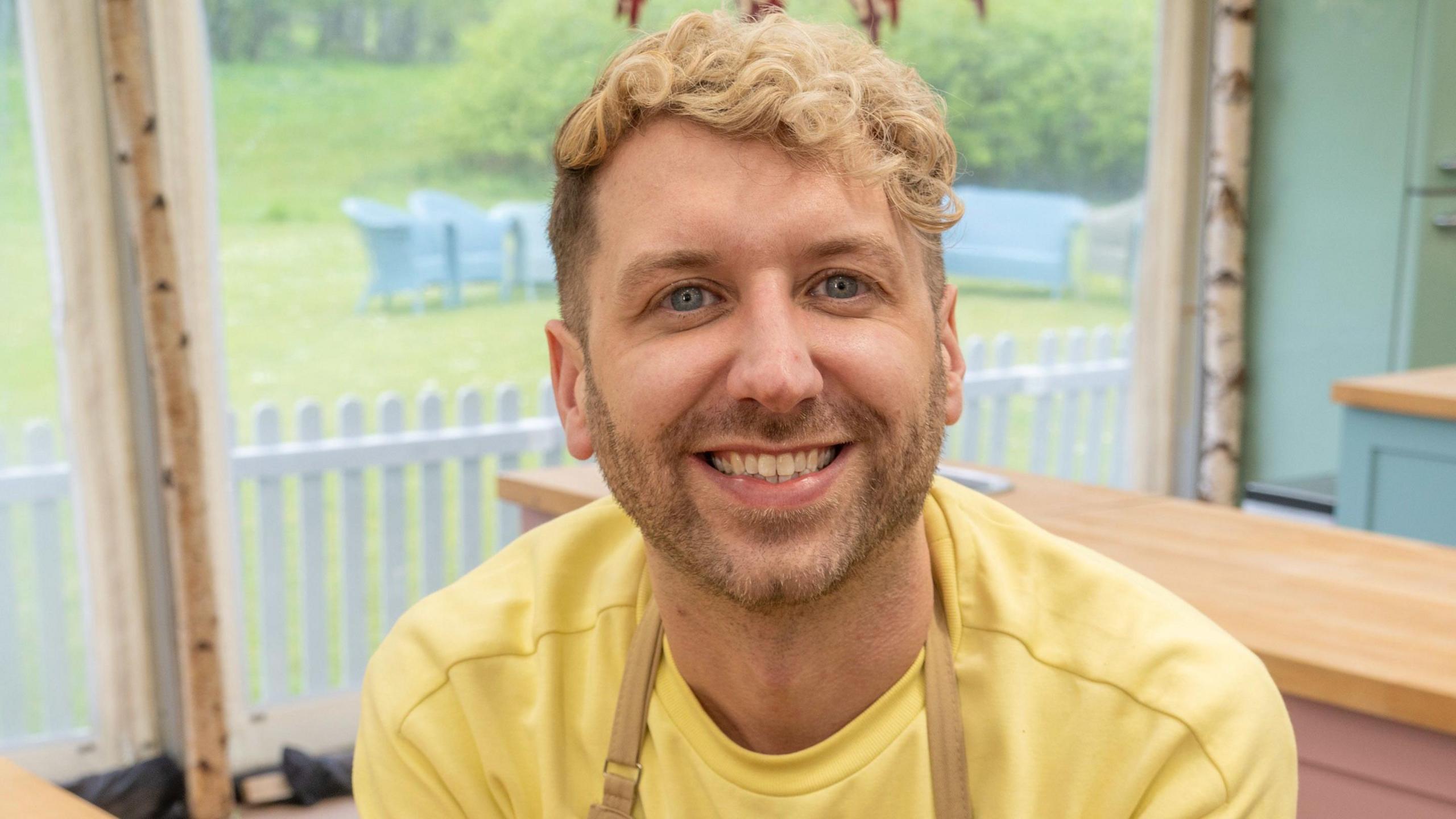 John Mincher, who has blonde hair and is wearing a yellow top, is in the Bake-off tent