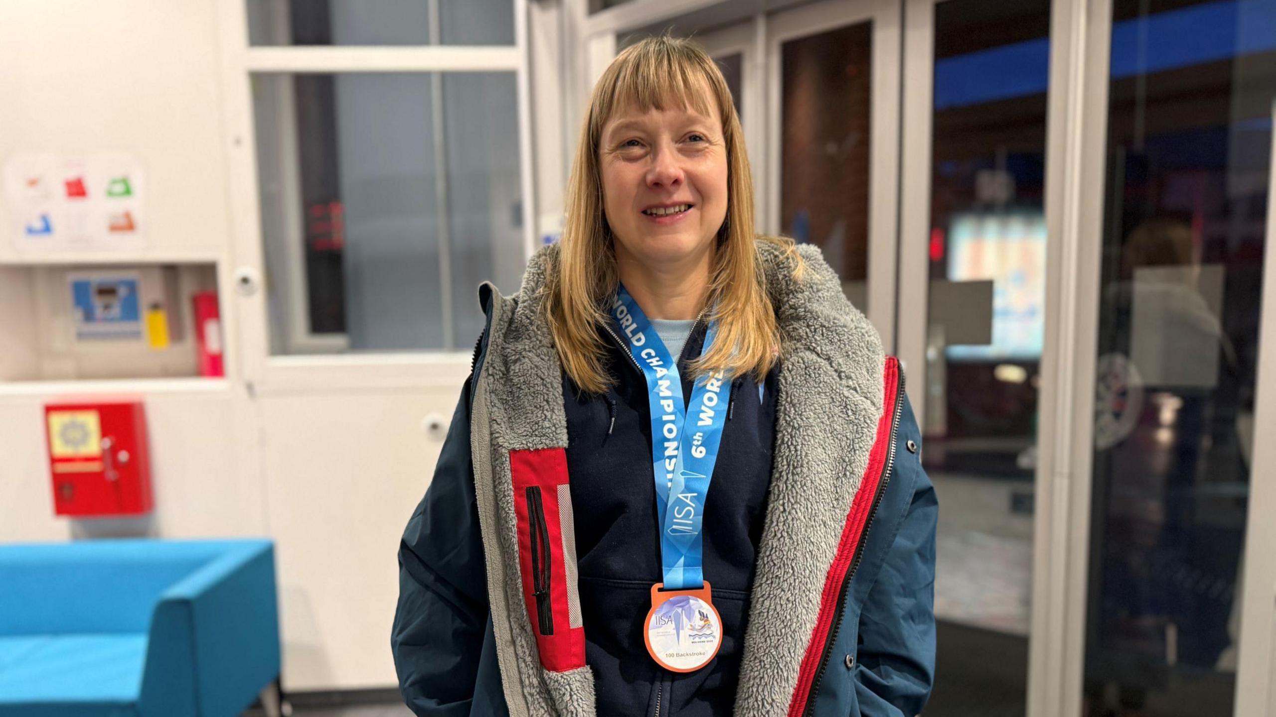 Amy Mellor smiling at the camera while wearing her medal around her neck. She has long blonde hair with a fringe and is wearing a padded outdoor jacket on top of a fleece. 