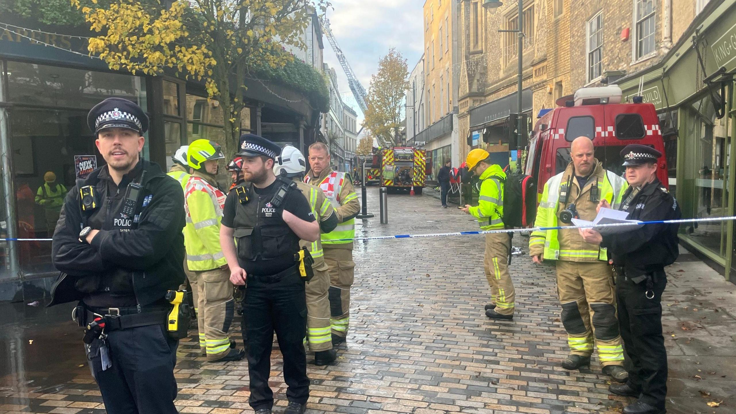 Police office and firefighters standing by blue and white police tape with fire vehicles and a ladder in the background.