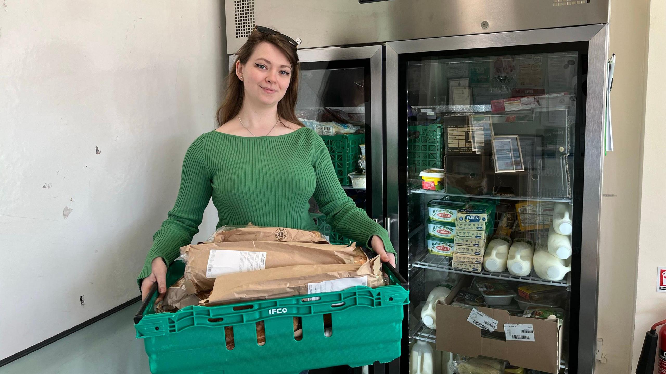 A woman in a green jumper holding a large tray in front of a fridge.