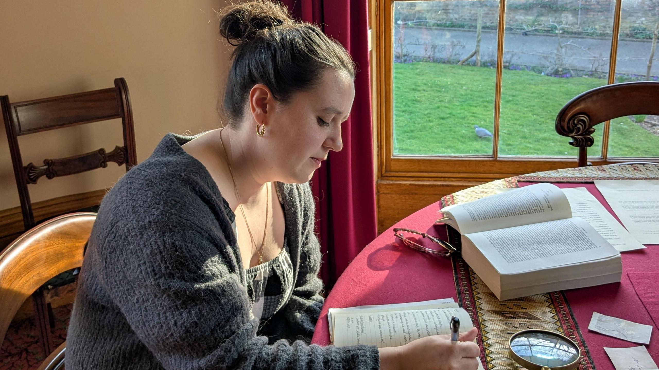 Georgia writing in her notebook on Gaskell's table, which has an open book and magnifiying glass on it, next to a window looking out on to the garden