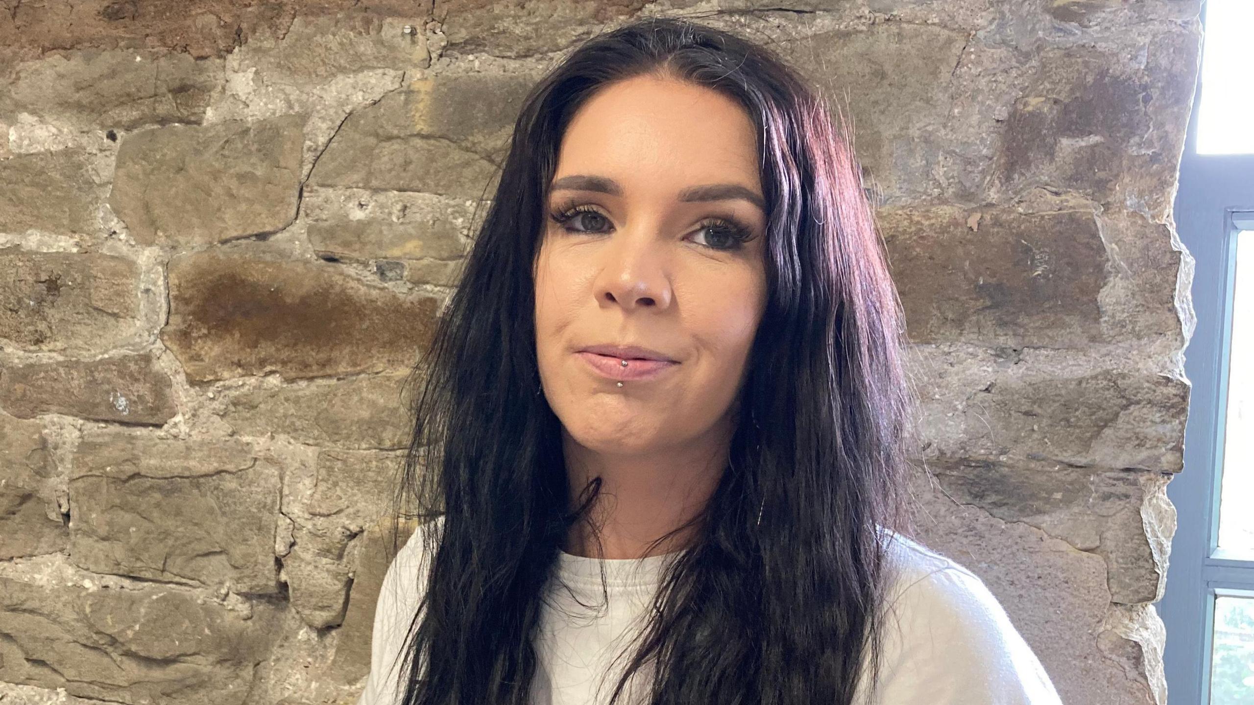 Key Crawford wearing a white t shirt with piercing on a lip standing in front of a stone wall next to a window. Key has long dark hair.