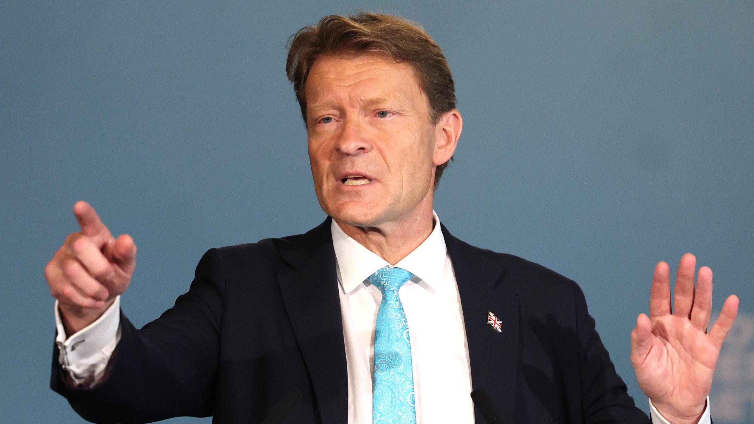 Richard Tice in front of a teal background, wearing a black suit, blue tie and white shirt, pointing into the foreground.