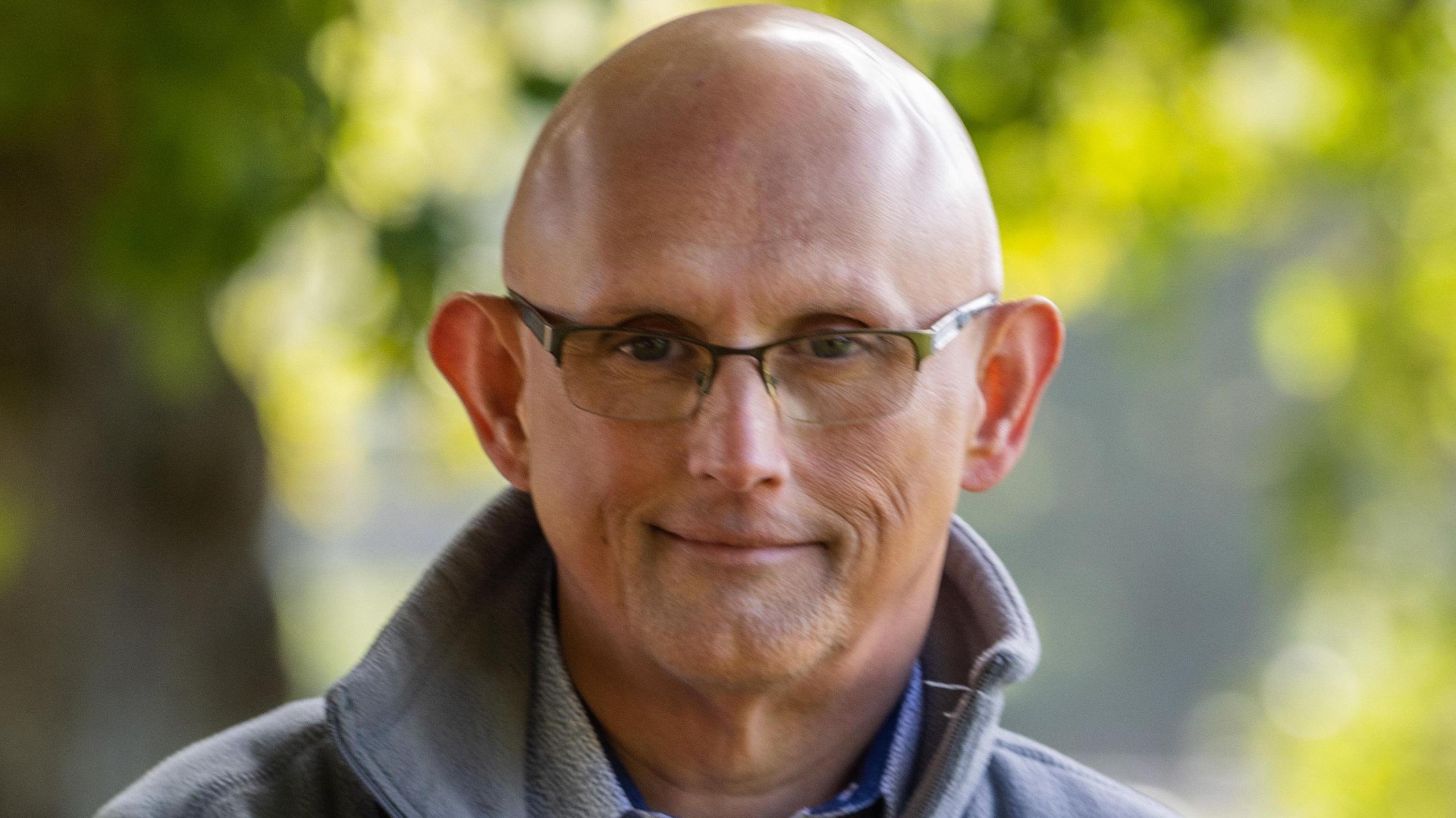 George Dunn is wearing a grey fleece and a blue shirt, he has half rimmed glasses and is pictured against a background of trees. 