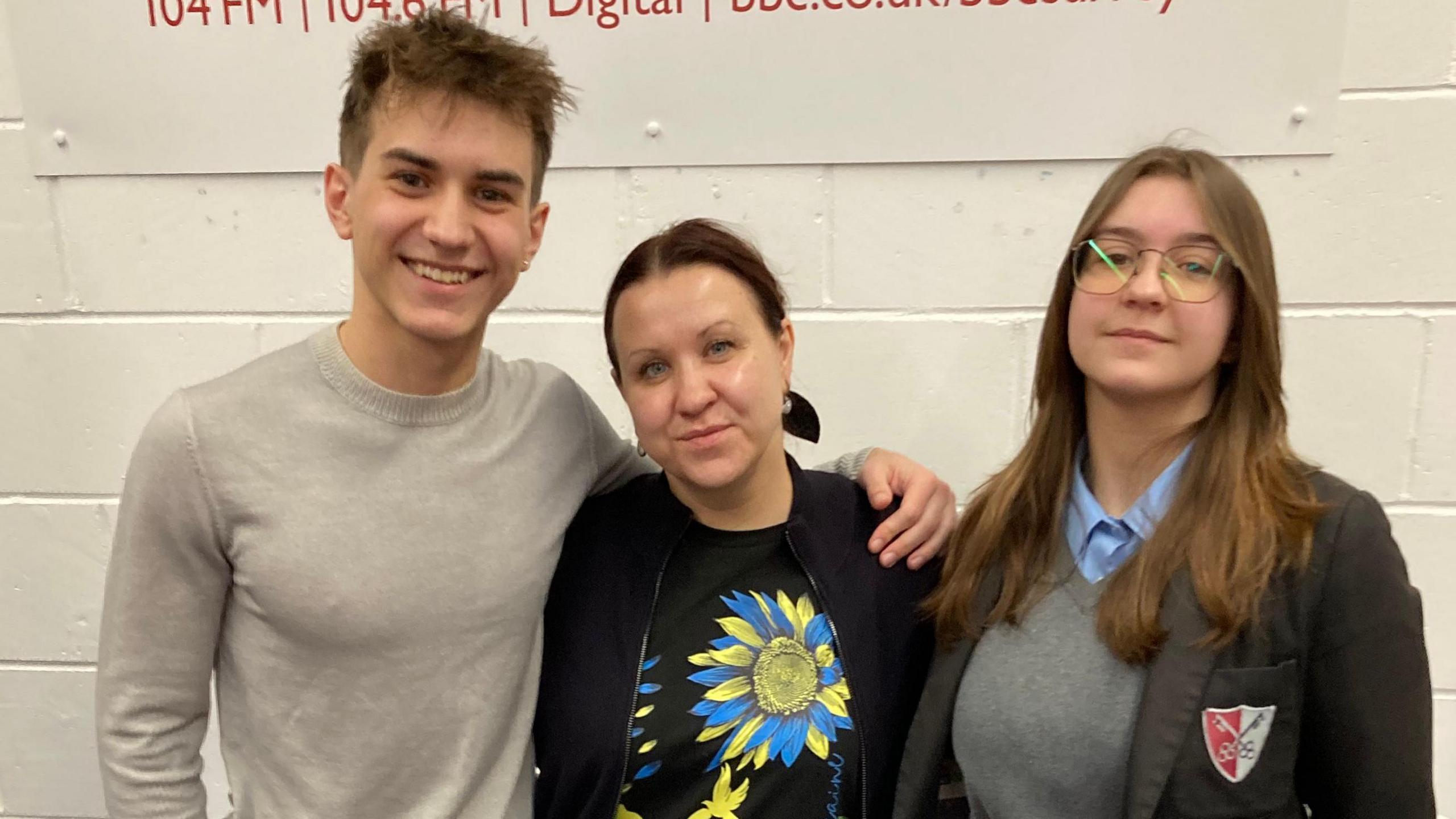 A mum and her teenage son and daughter stood in front of a white wall. All three are looking directly at the camera. 