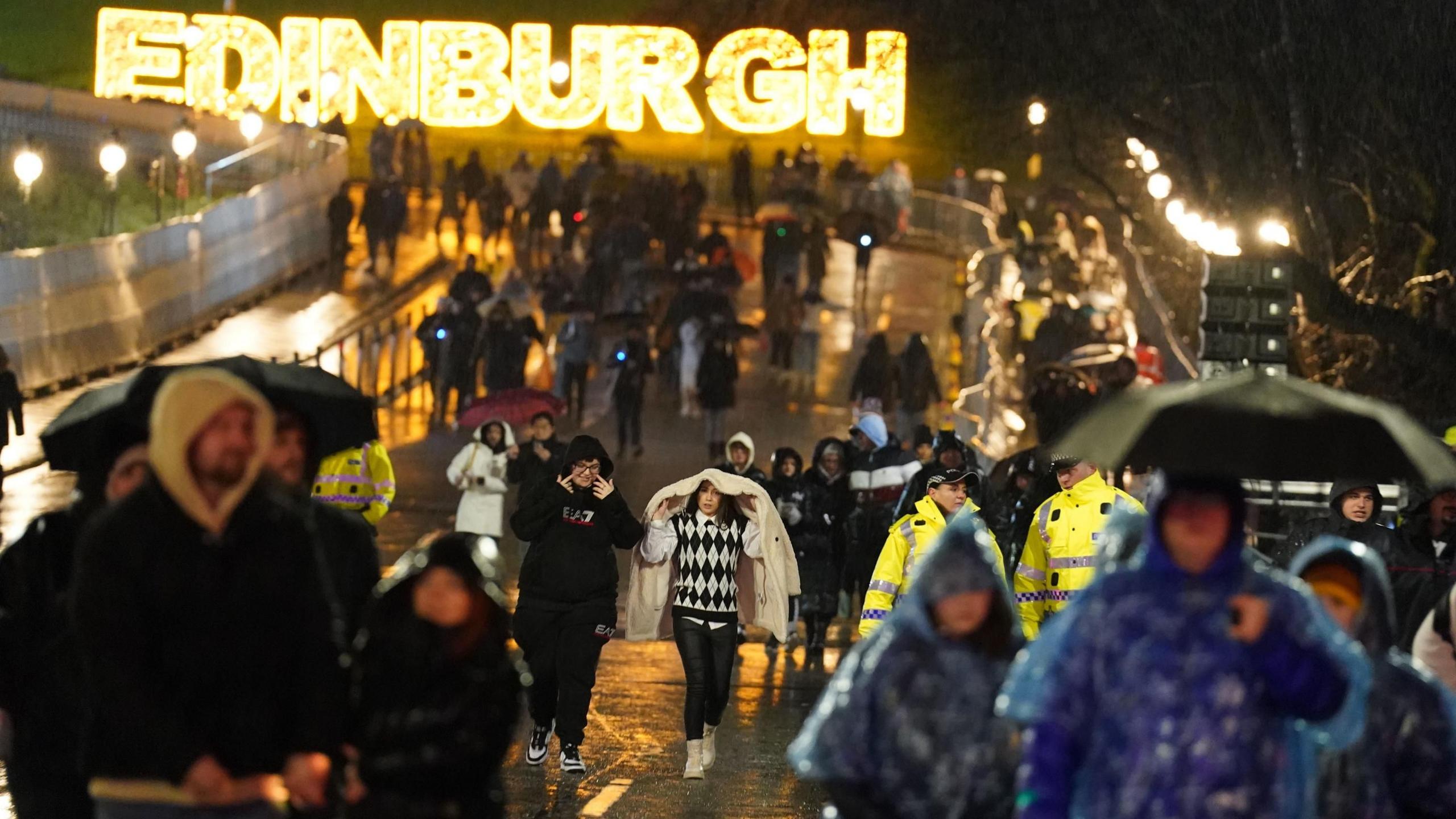 People walking in the rain in Edinburgh on Hogmanay in 2022. There is an illuminated Edinburgh sign in the background. The street is busy with people holding umbrellas or with hoods or jackets over their heads.