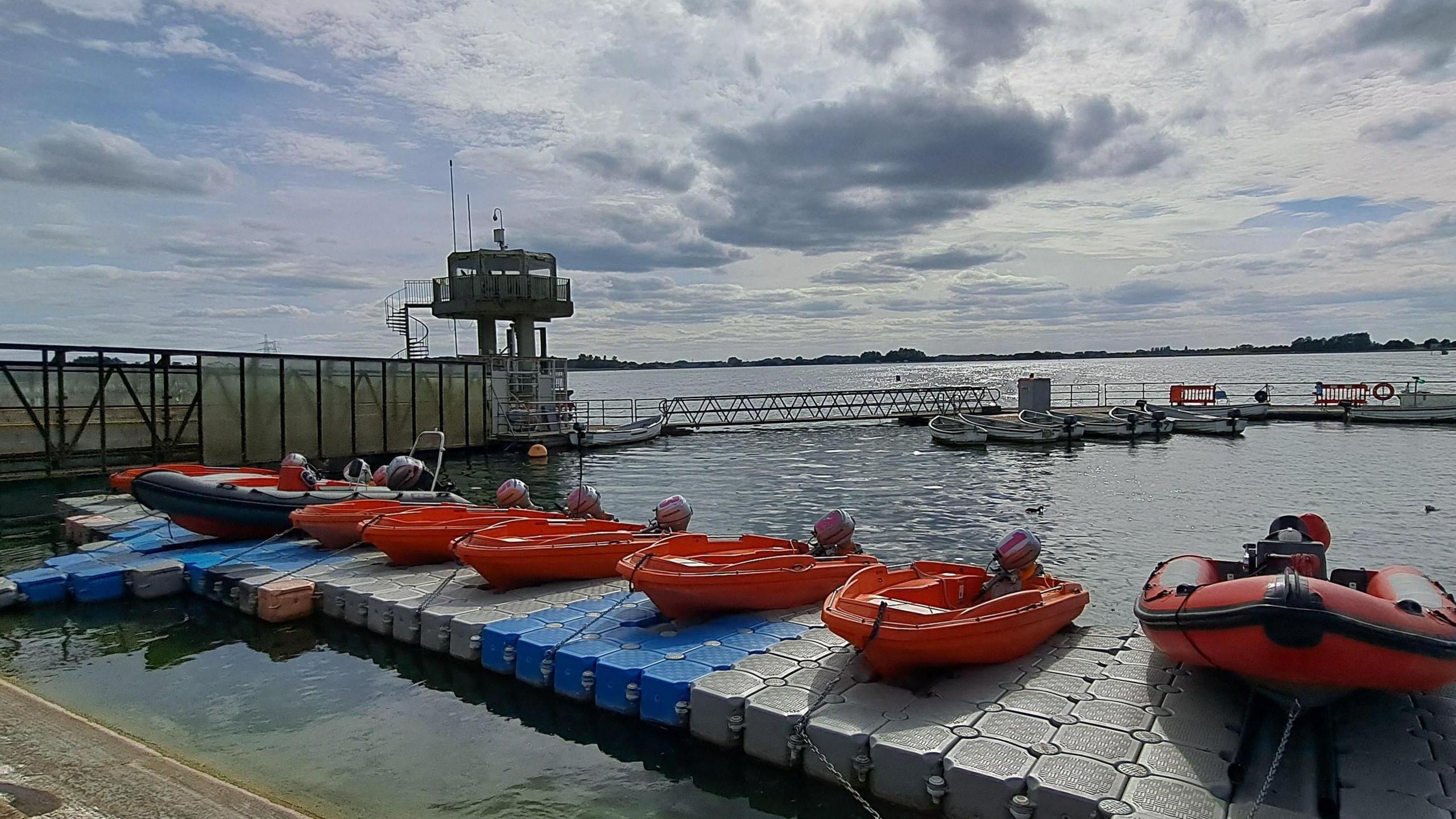 A cloudy bright sky with blue patches gives way to shimmering water of a reservoir. Several orange boats sit on a pontoon in the foreground.