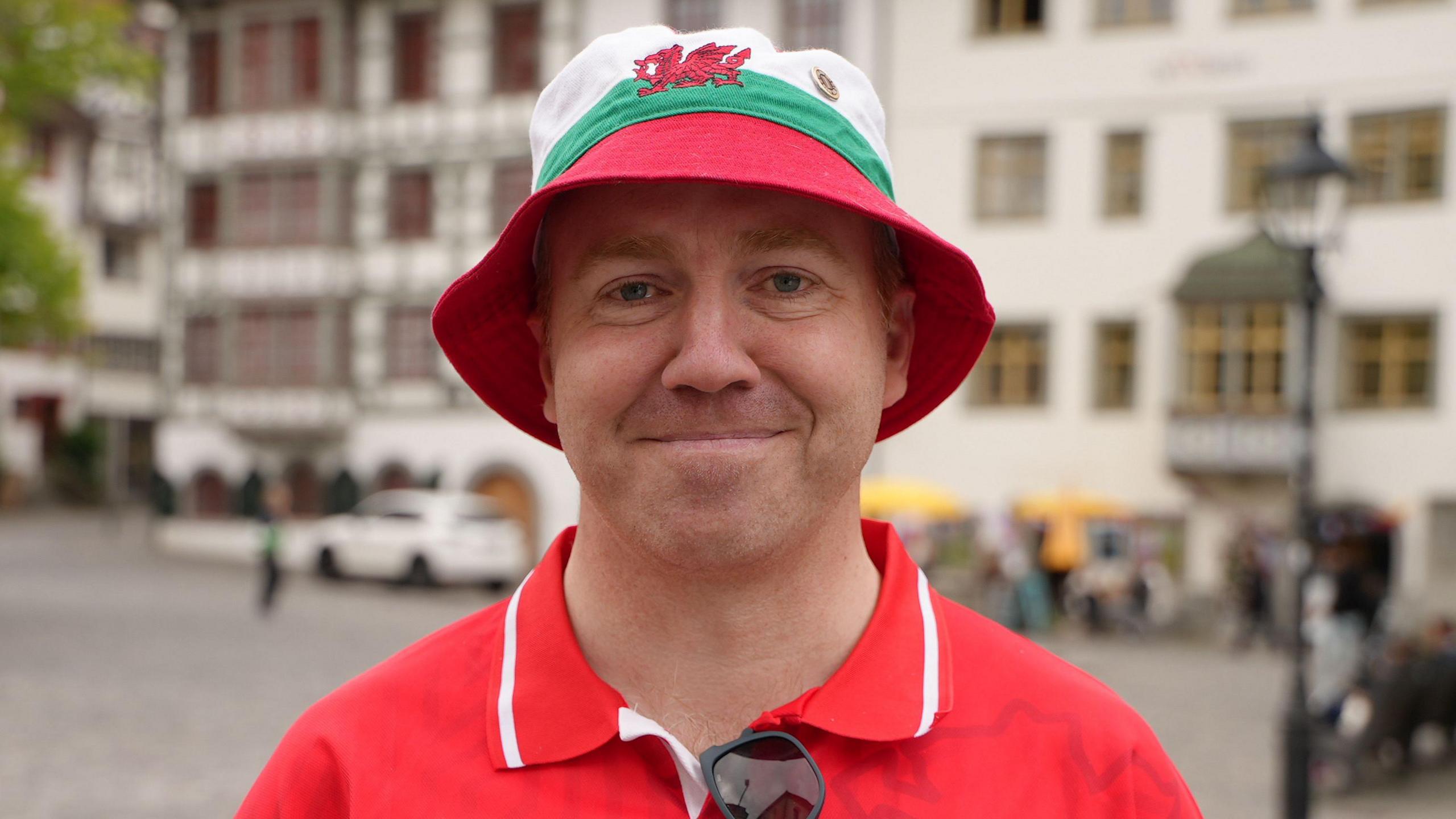 Gareth Reid smiling at the camera. He is wearing a red and white Wales shirt and a red, white and green bucket hat with the Welsh dragon at the front. He is standing on a street with yellow cars behind him.