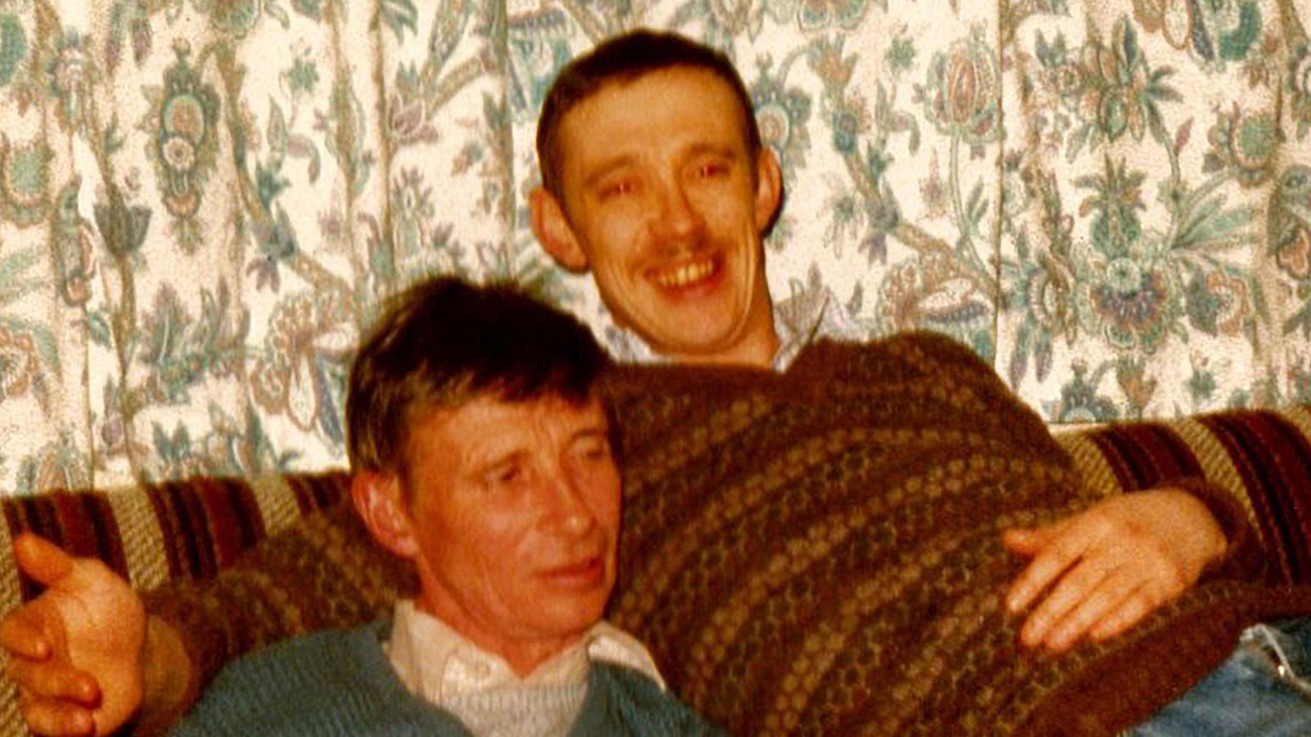 Ean Coutts in younger years. He has short hair and a moustache and is smiling at the camera. He is wearing a brown, patterned V-neck jumper and jeans. He is sitting on a brown, striped sofa and has his arm around another man  who is wearing a green jumper over a shirt.