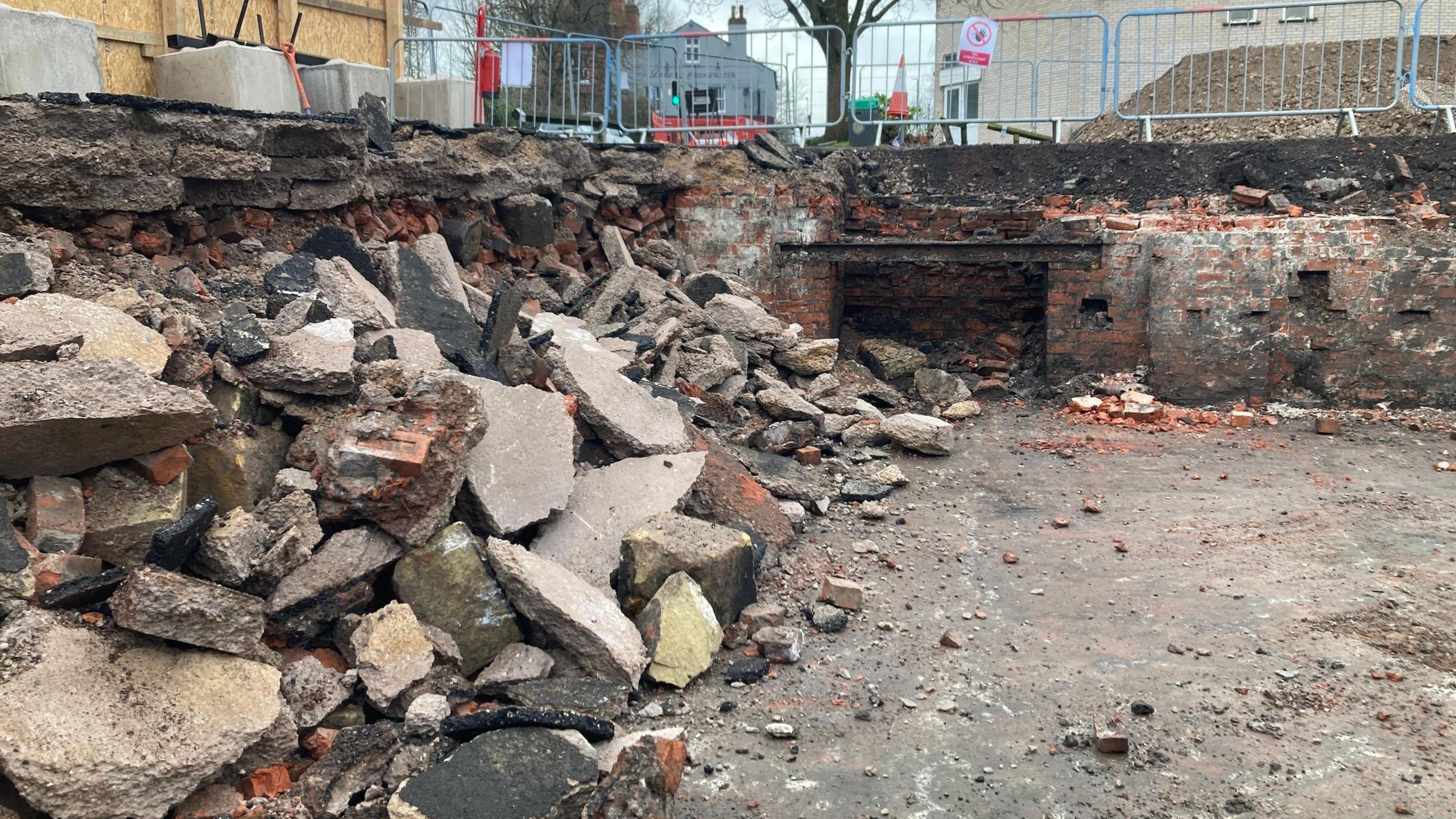 An excavation site - to the left there is a large pile of rubble made up of large broken pieces of brick and concrete. In the background of the image is a brick wall which has a thick layer of asphalt on top of it